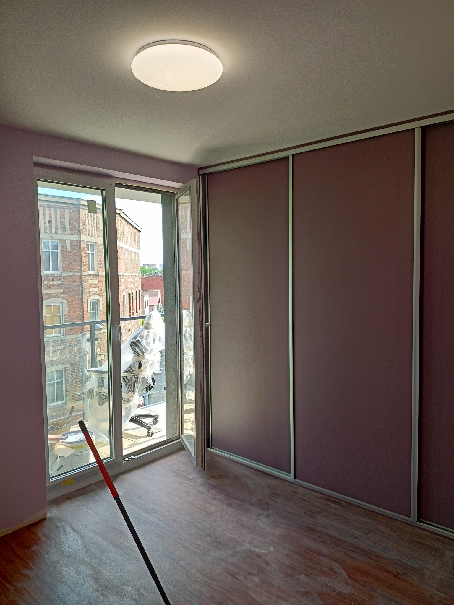 Pokój po remoncie: widok na zewnątrz przez otwarte okno, częściowo zasłonięty folią meblową, brązowa podłoga, przesuwne drzwi szafy w kolorze burgundowym, widoczny kij od mopa.