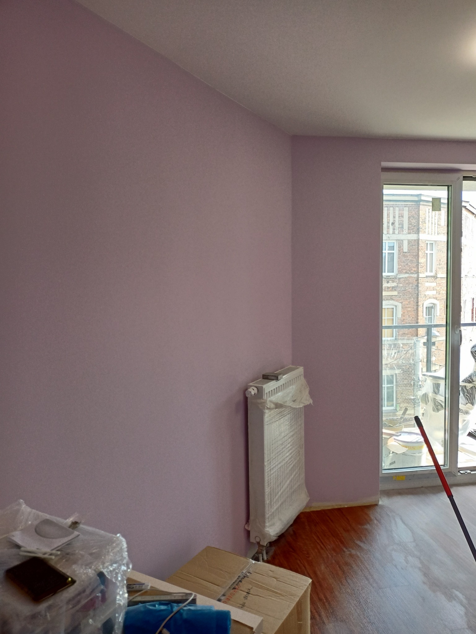 Pokój po malowaniu ścian na liliowo, z zabezpieczonym grzejnikiem i widocznym fragmentem podłogi oraz okna z widokiem na budynek z cegły.