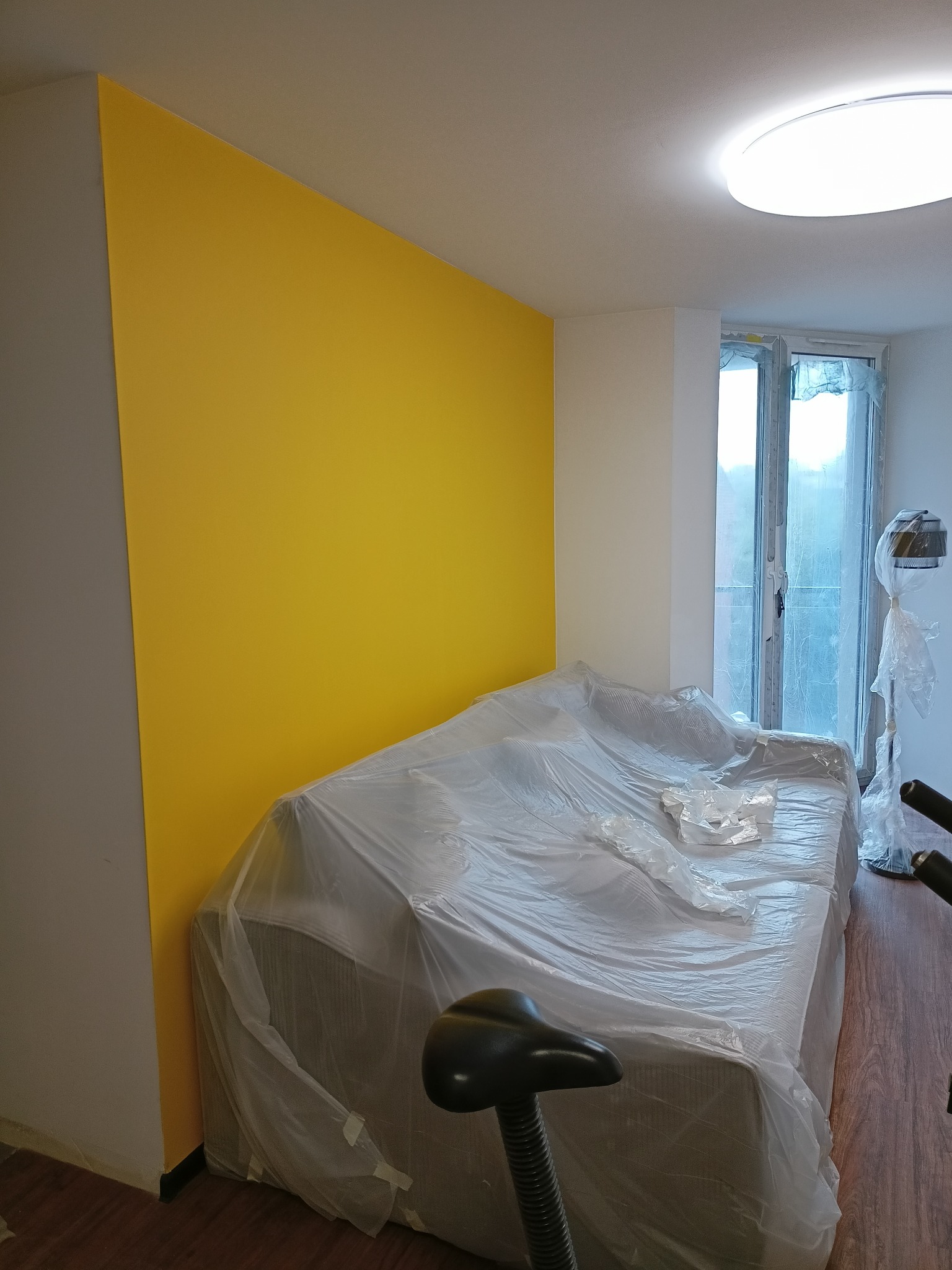 Żółta ściana po świeżym pomalowaniu, zabezpieczona folią sofa, widoczny rower stacjonarny i okno balkonowe.