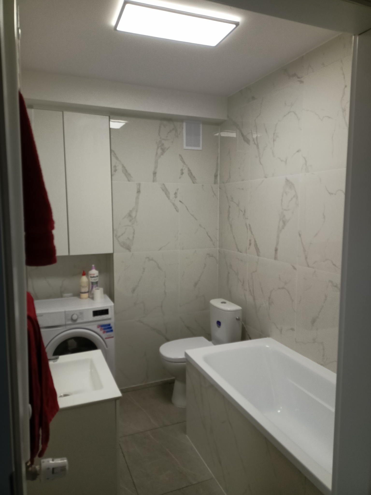 Nowoczesna łazienka wykończona płytkami imitującymi marmur, z białą wanną, toaletą, pralką i szafkami, oświetlona jasnym, prostokątnym panelem LED.