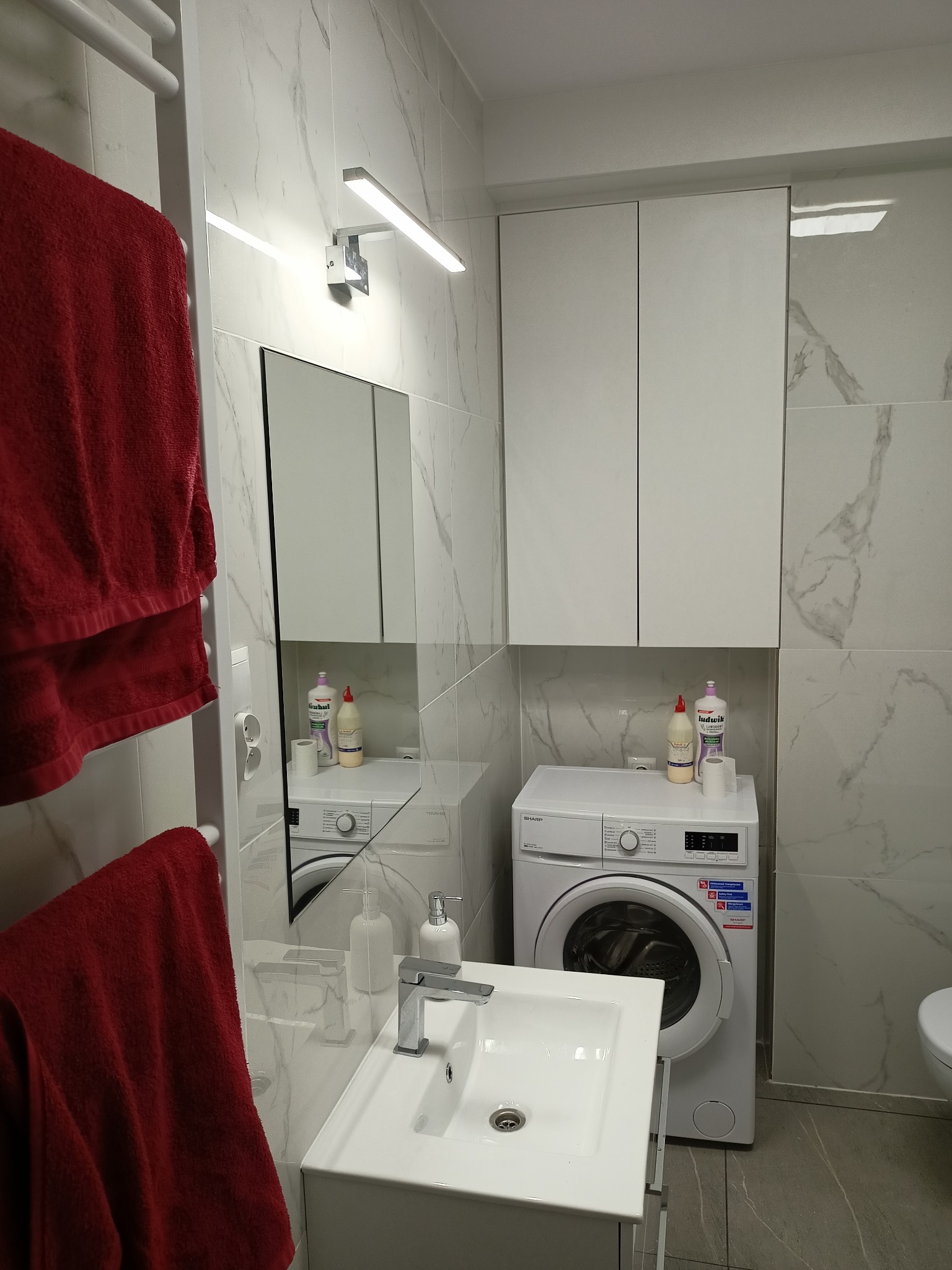 Nowoczesna łazienka z marmurowymi płytkami, białą umywalką z szafką, pralką i wiszącymi szafkami, oświetlona listwą LED, z czerwonym ręcznikiem na grzejniku.