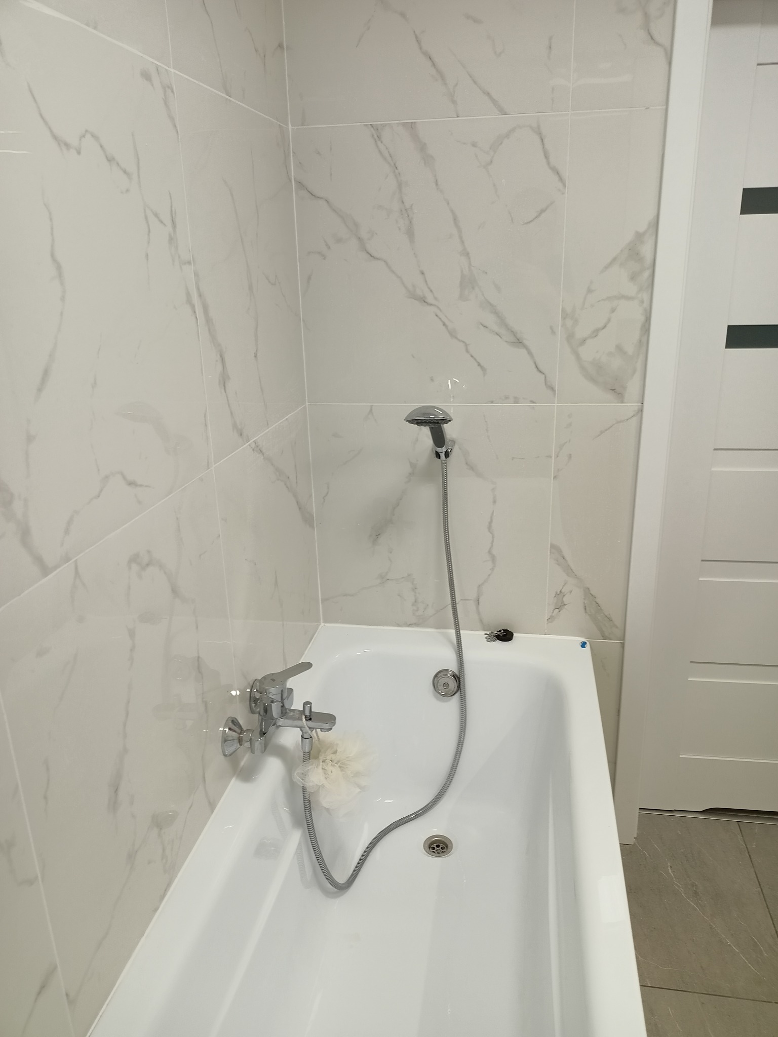Biała wanna z zamontowaną baterią i słuchawką prysznicową na tle ściany wyłożonej płytkami imitującymi marmur, widoczny fragment białych drzwi z czarnymi wstawkami i szarej podłogi.
