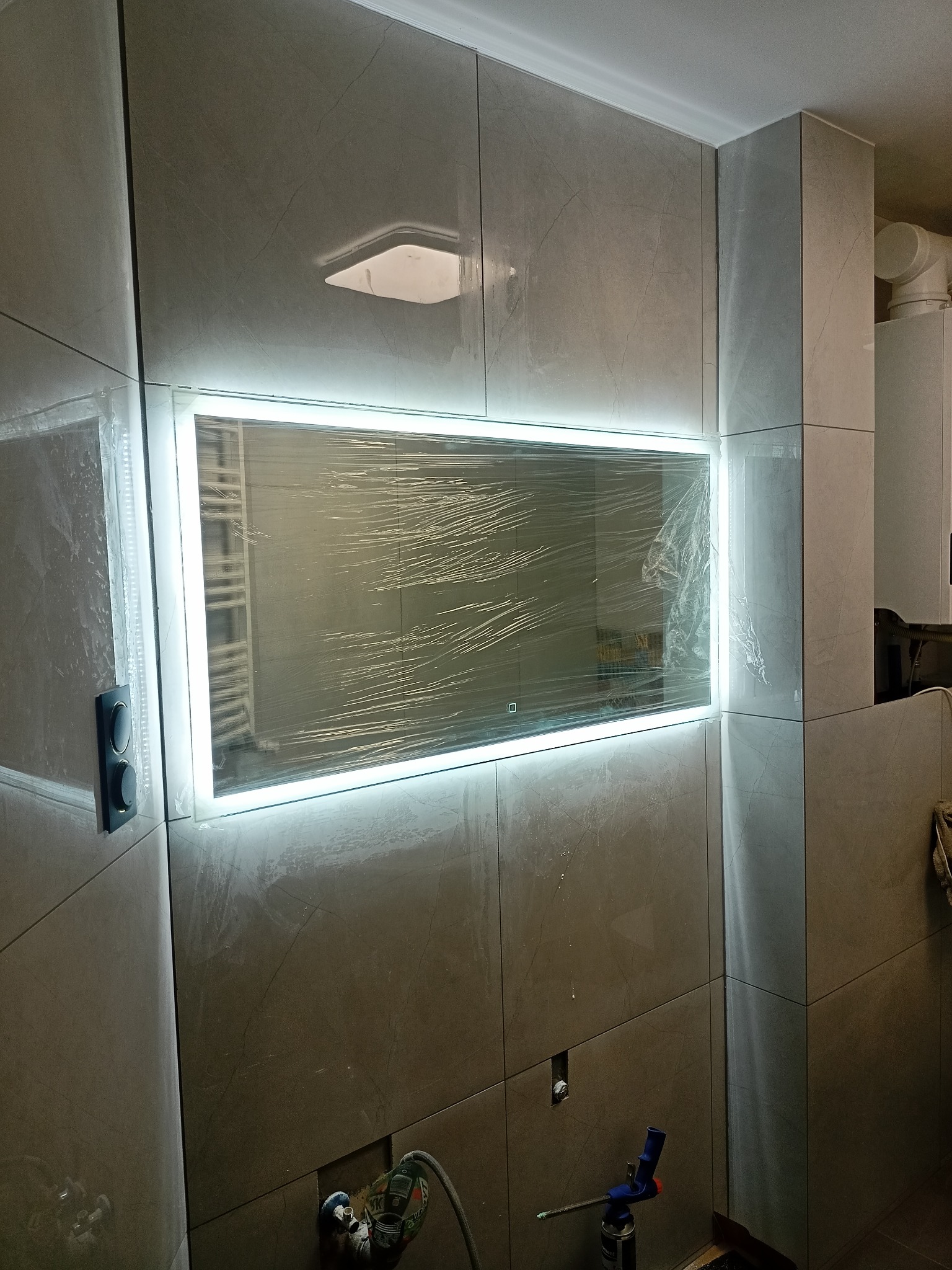 Świeżo wyremontowana łazienka z podświetlanym lustrem w folii ochronnej, płytki imitujące marmur na ścianach, widoczne narzędzia i instalacja elektryczna.