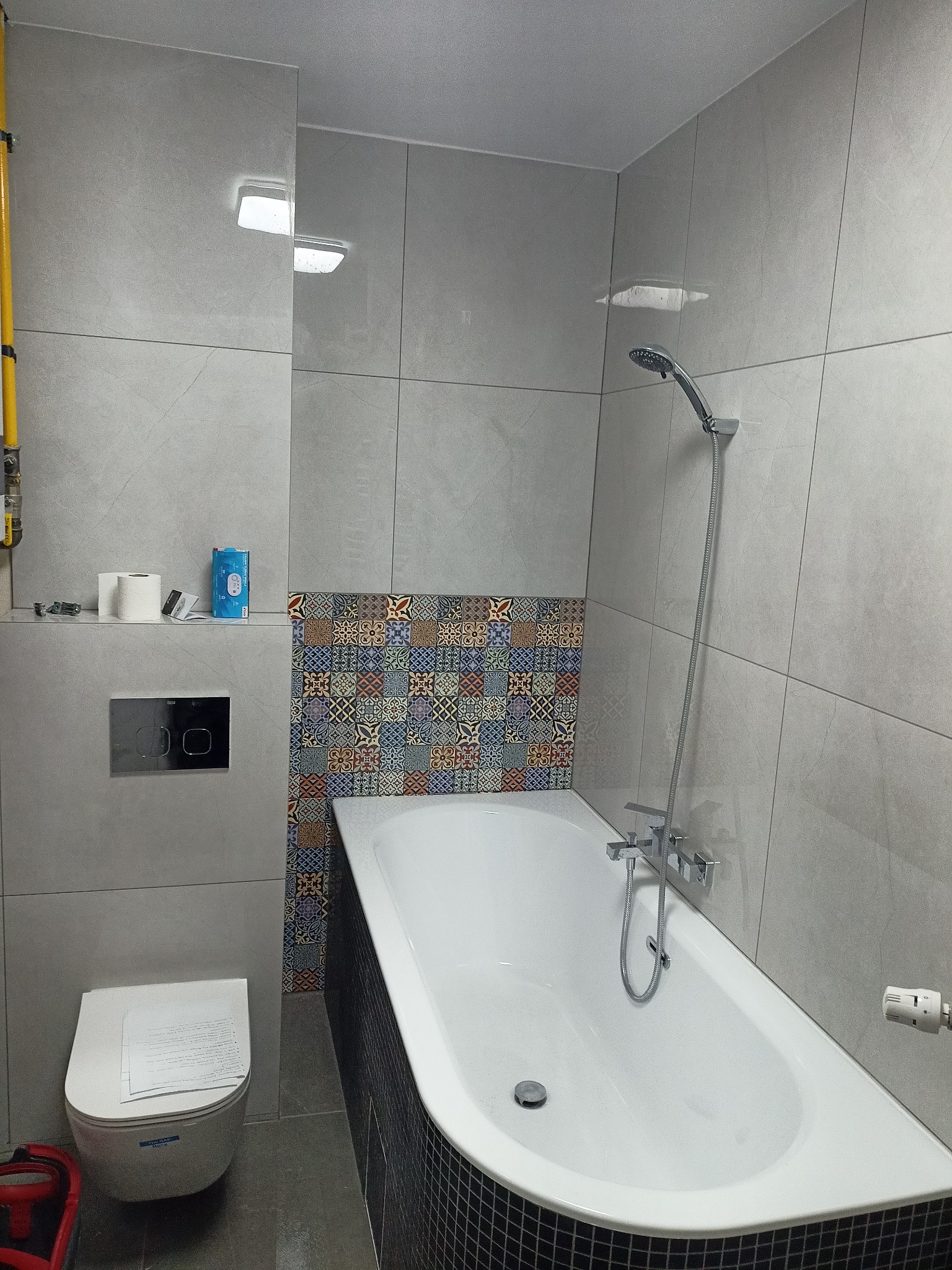 Wykończona łazienka z wanną obudowaną czarną mozaiką, szare płytki na ścianach, dekoracyjny pas płytek nad wanną, podwieszana toaleta z dokumentami na desce, żółta instalacja gazowa widoczna po...