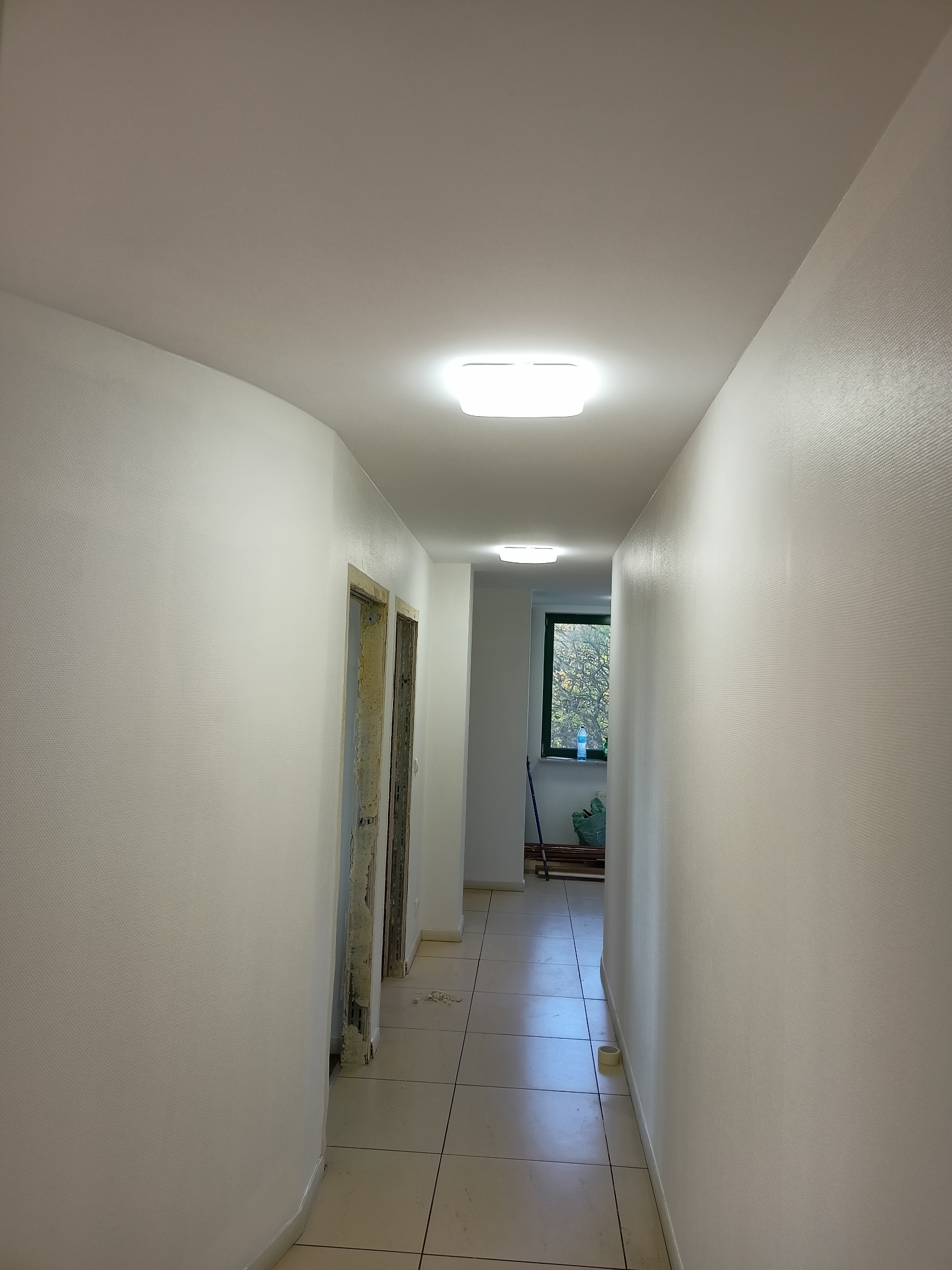 Korytarz po świeżym malowaniu na biało, widoczne odnowione ściany z drobną fakturą, nowe oświetlenie sufitowe LED i przygotowane do montażu ościeżnice drzwiowe.