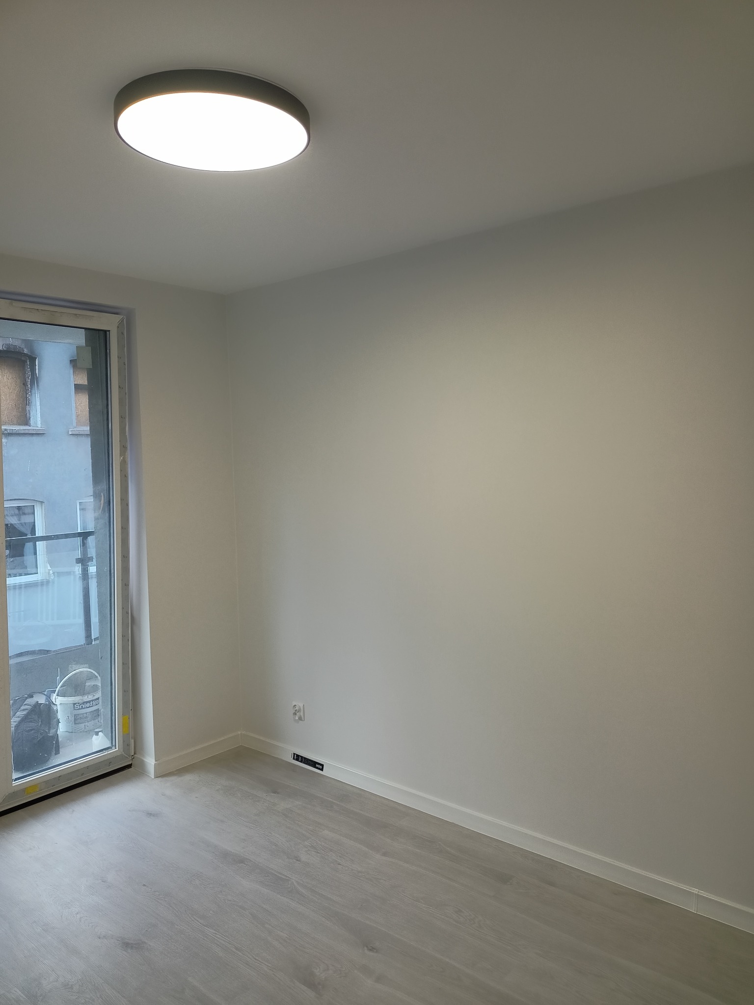 Wnętrze pokoju po remoncie: świeżo pomalowane ściany w odcieniach bieli, jasna podłoga imitująca drewno, okno balkonowe z widokiem na zewnątrz, minimalistyczna lampa sufitowa.