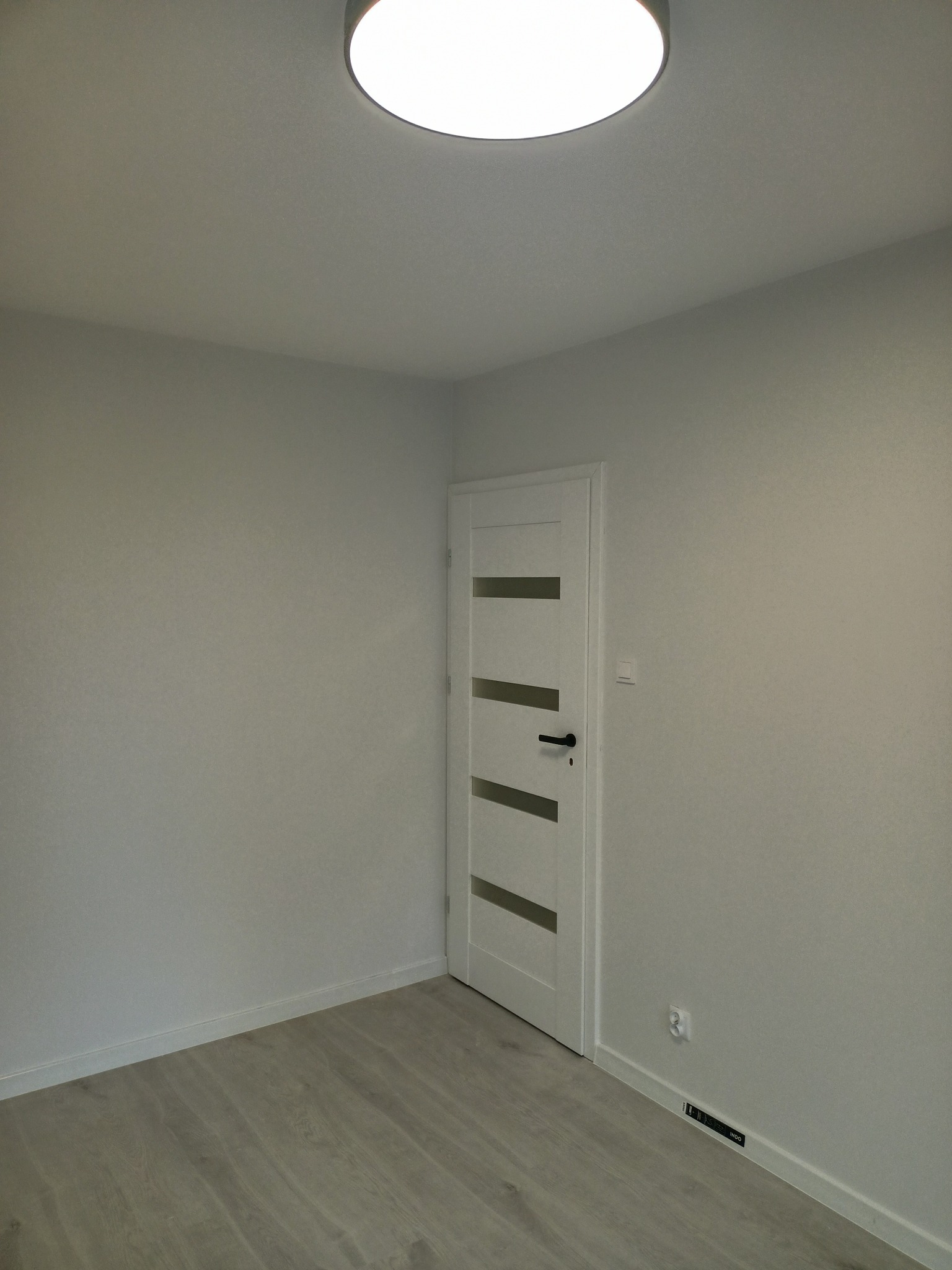 Pomalowane na szaro wnętrze pokoju z drewnopodobną podłogą, białe drzwi z poziomymi przeszkleniami i okrągła lampa sufitowa.