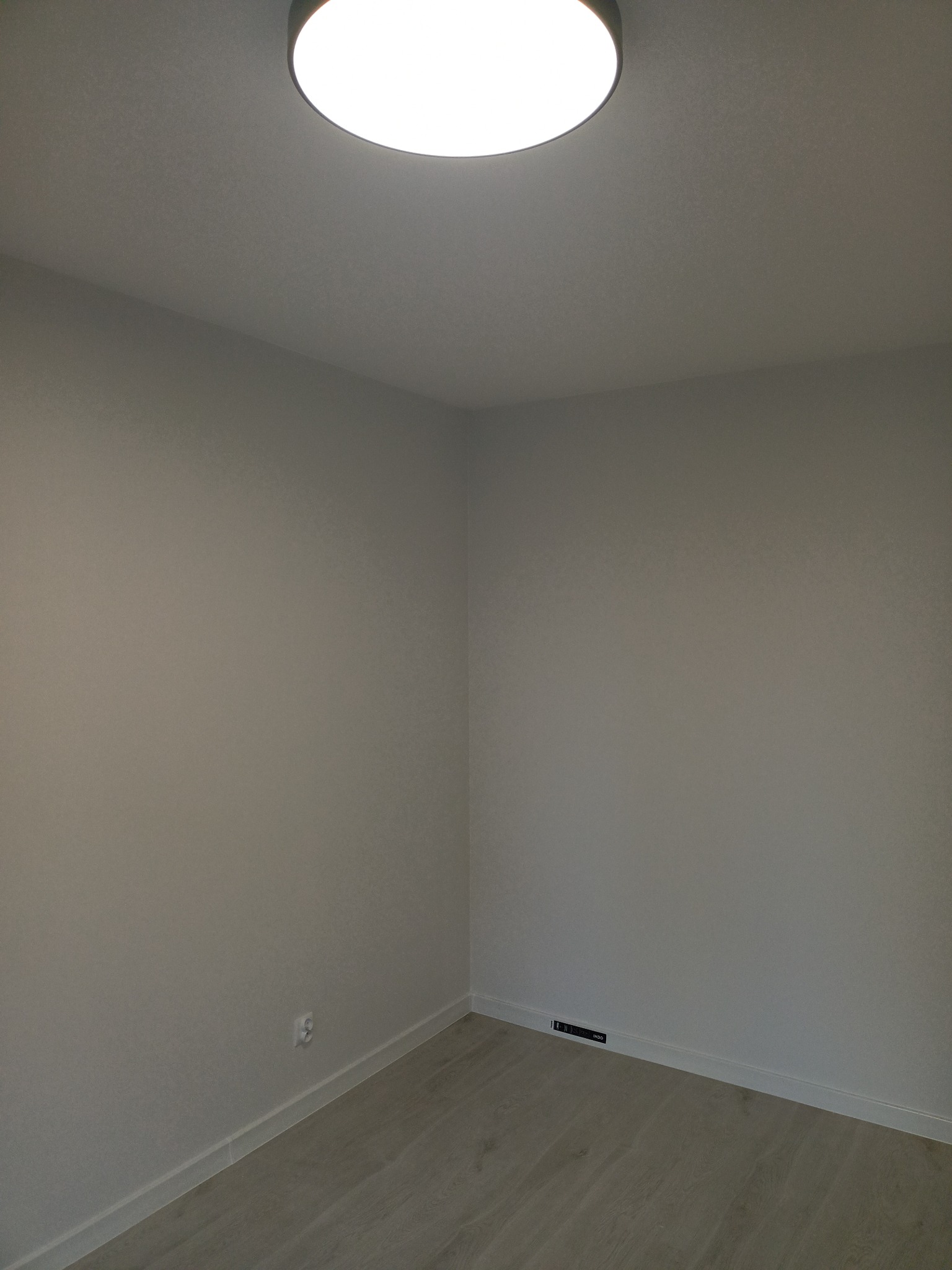 Pomalowane na szaro wnętrze pokoju z panelami podłogowymi i okrągłą lampą sufitową, widoczny kontakt elektryczny przy podłodze oraz kratka wentylacyjna.