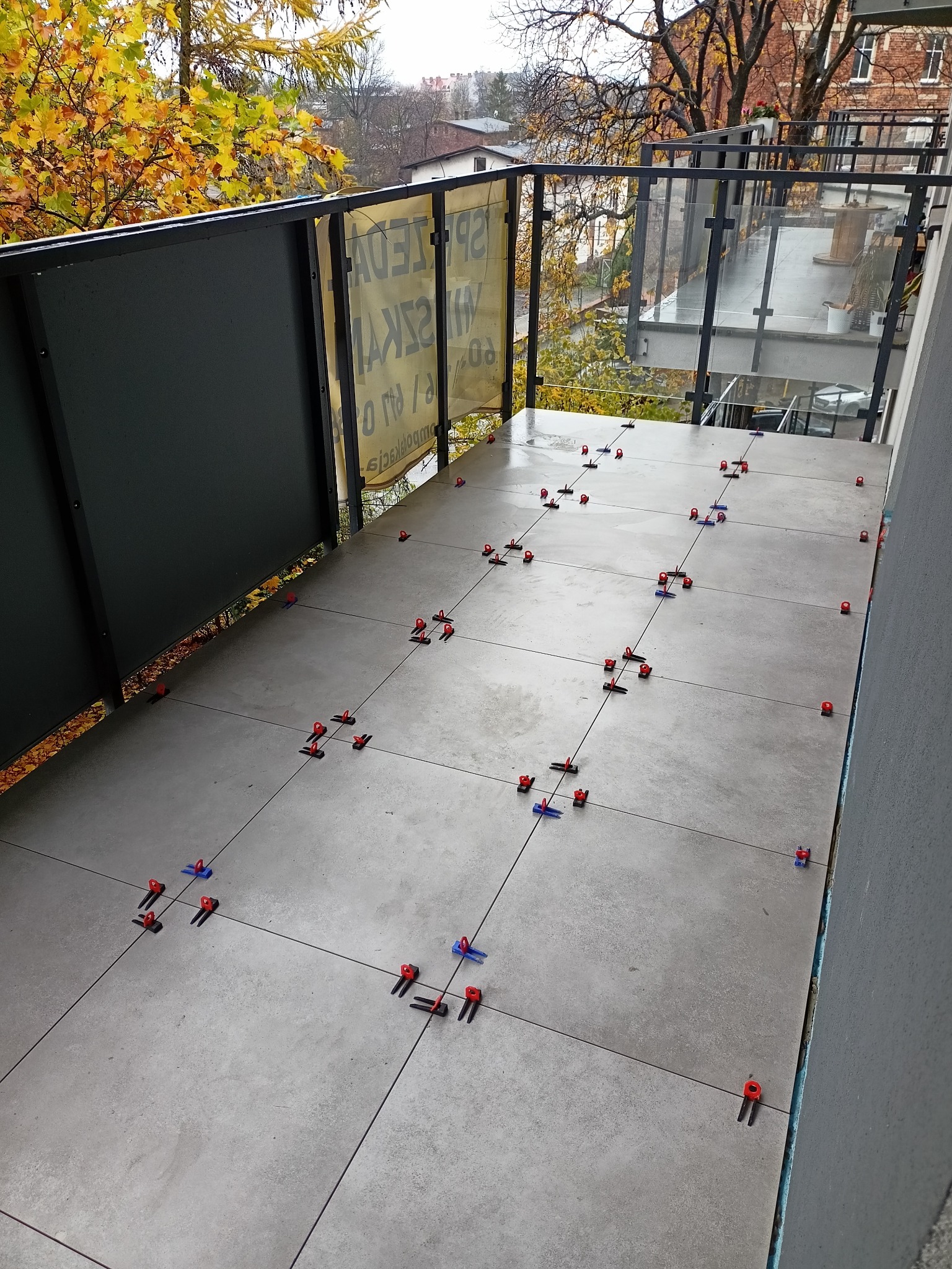 Szare płytki ułożone na balkonie z użyciem systemu poziomowania, widoczne jesienne drzewa za szklaną balustradą.