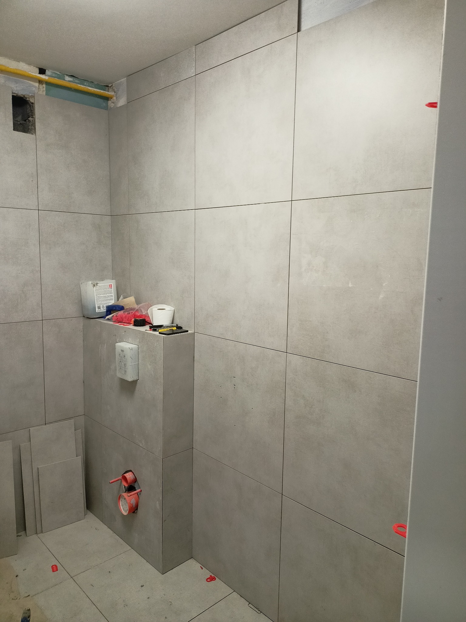 Ściana w łazience w trakcie wykańczania, widoczne szare płytki ceramiczne z czerwonymi krzyżykami dystansowymi, niezabudowana instalacja sanitarna, narzędzia i materiały budowlane na półce.