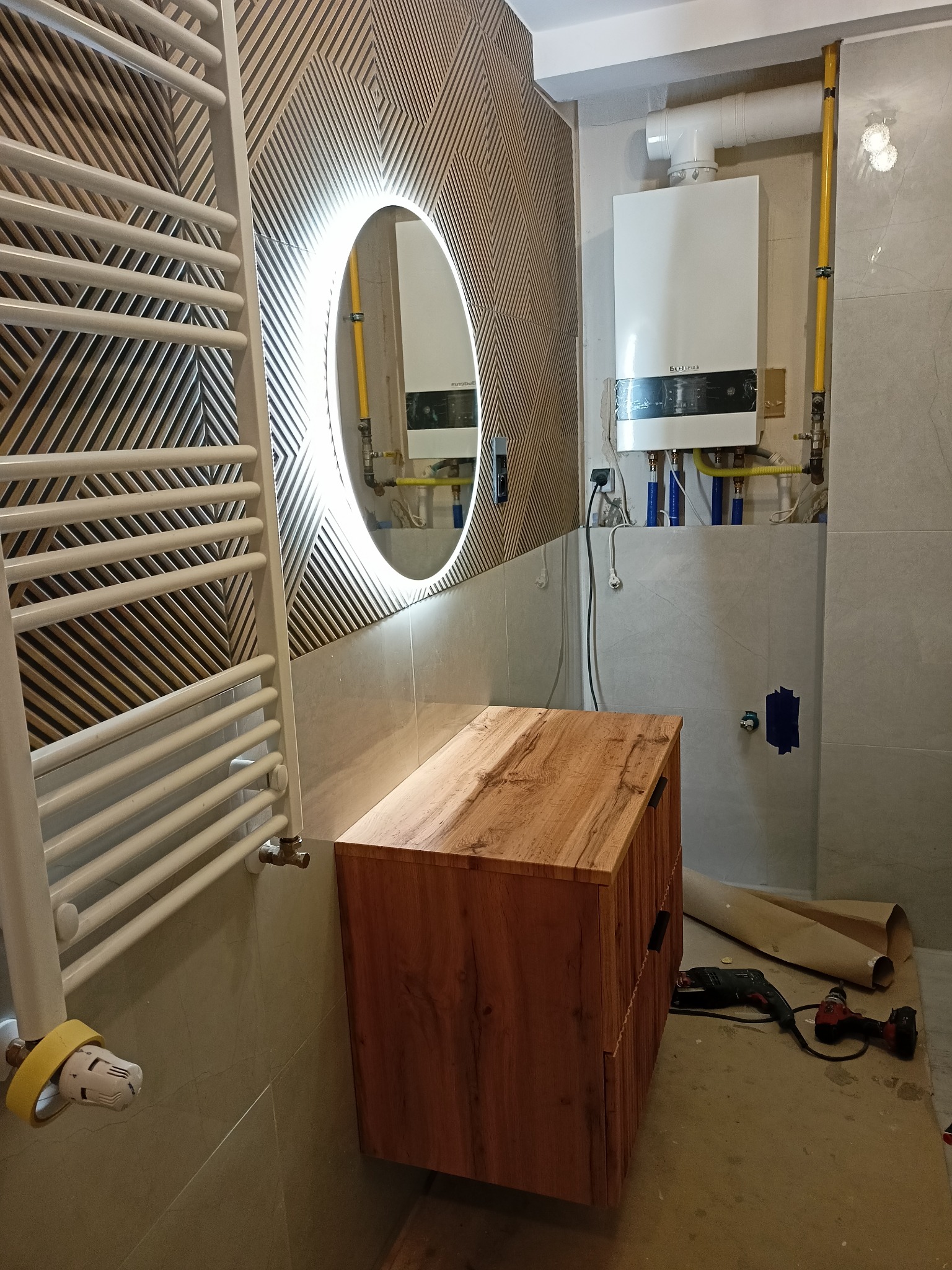 Nowoczesna łazienka w trakcie remontu; widoczna szafka pod umywalkę z drewnianym wykończeniem, grzejnik drabinkowy, okrągłe lustro z podświetleniem LED oraz piec gazowy na ścianie; na podłodze...