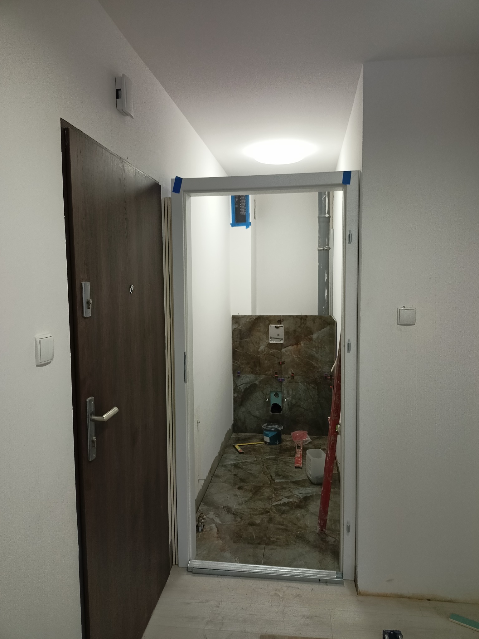 Wnętrze łazienki w trakcie remontu: widoczne płytki imitujące marmur na ścianie, rury, oraz narzędzia i materiały budowlane, widok z korytarza przez ościeżnicę bez drzwi.