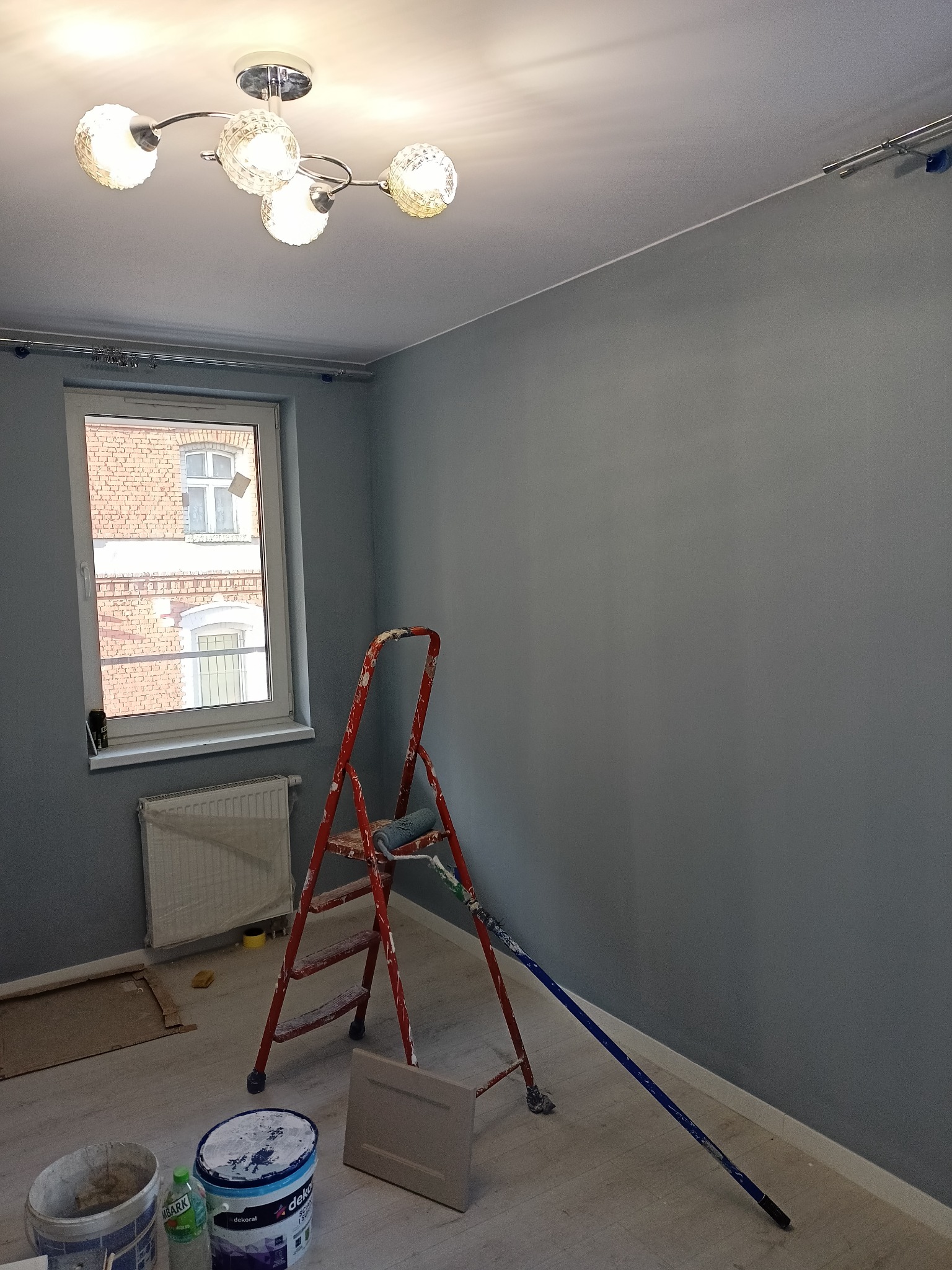 Pokój po świeżym pomalowaniu na szaro, z widoczną drabiną, wałkiem malarskim, kuwetą i puszkami farb, z widokiem przez okno na budynek z czerwonej cegły.