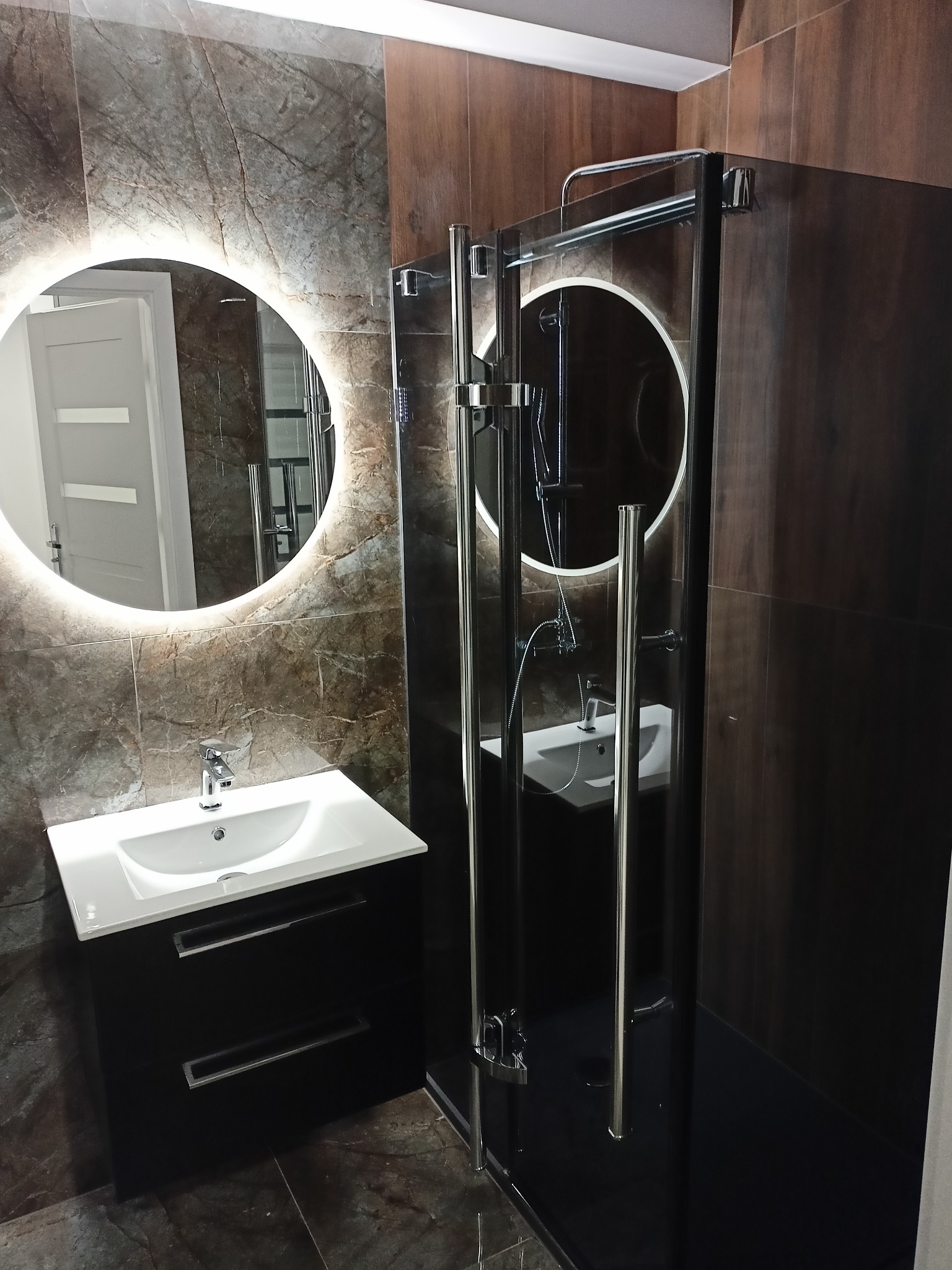 Nowoczesna łazienka z kabiną prysznicową typu walk-in, umywalką nablatową na czarnej szafce z dwoma szufladami i okrągłym lustrem z podświetleniem LED.