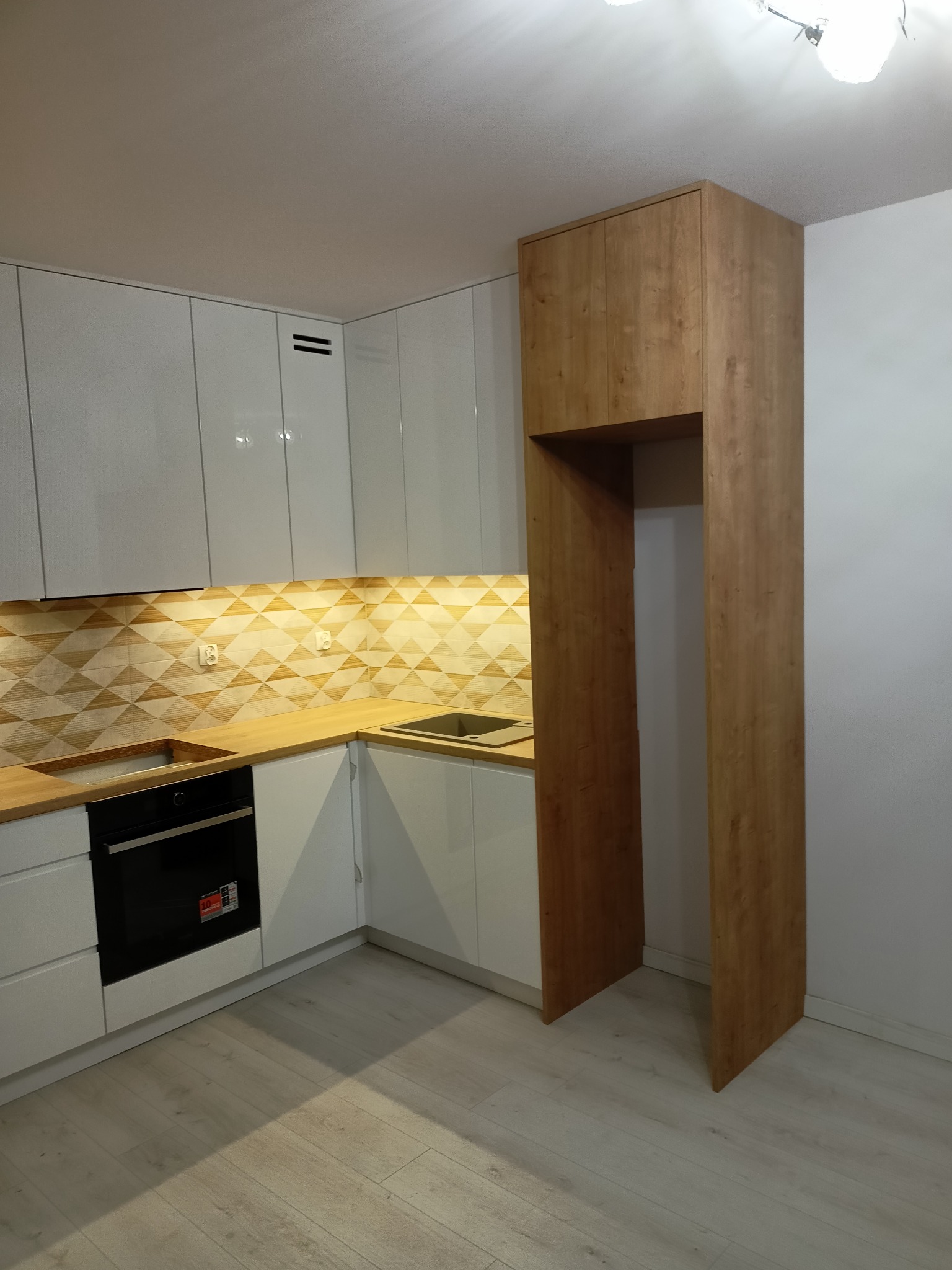 Nowoczesna kuchnia w Zabrzu z białymi, lakierowanymi szafkami, drewnianym blatem i geometryczną mozaiką nad blatem, z zabudowaną obudową na lodówkę z drewna. Widoczny piekarnik w zabudowie i jasna...