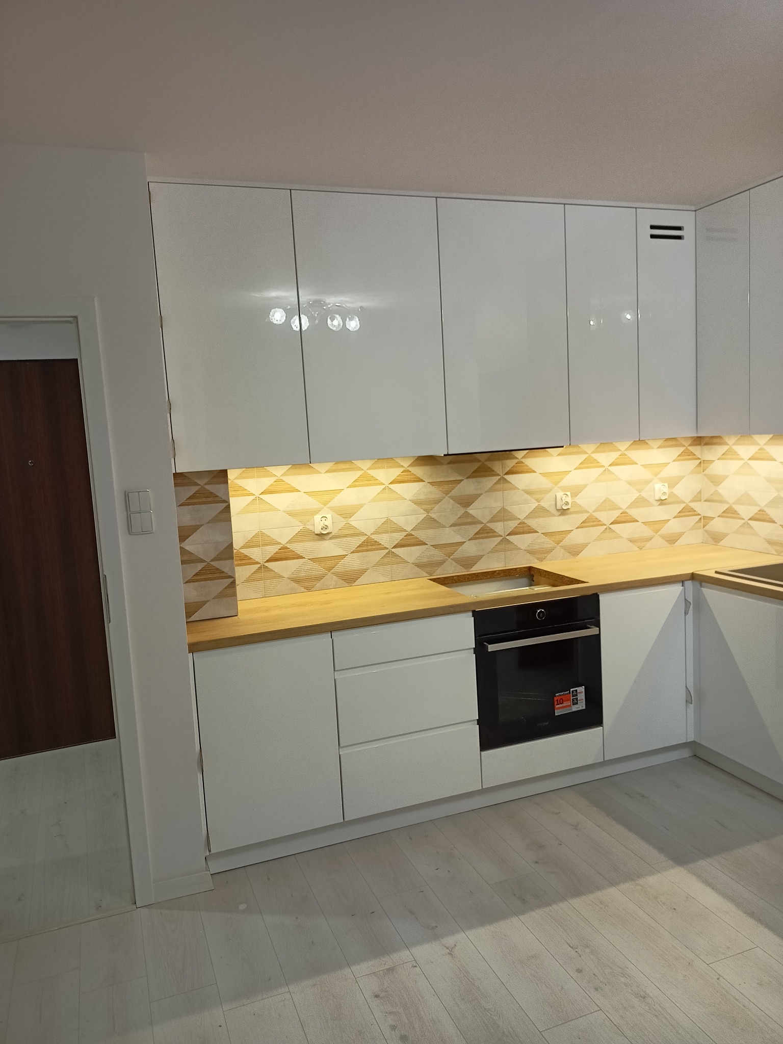 Biała, nowoczesna zabudowa kuchenna z drewnianym blatem i geometryczną mozaiką na ścianie, oświetlona punktowymi lampami podszafkowymi, z widocznym piekarnikiem w zabudowie.