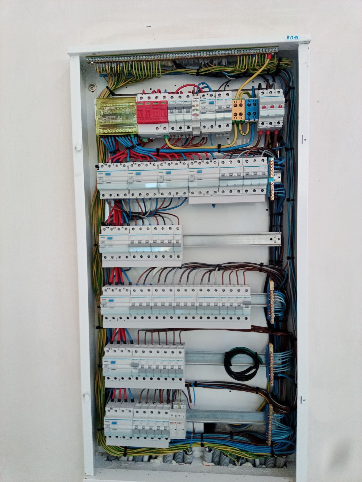 Rozdzielnica elektryczna marki Eaton w białej obudowie, z widocznymi bezpiecznikami, przewodami w różnych kolorach i listwami zaciskowymi, zamontowana na jasnej ścianie.