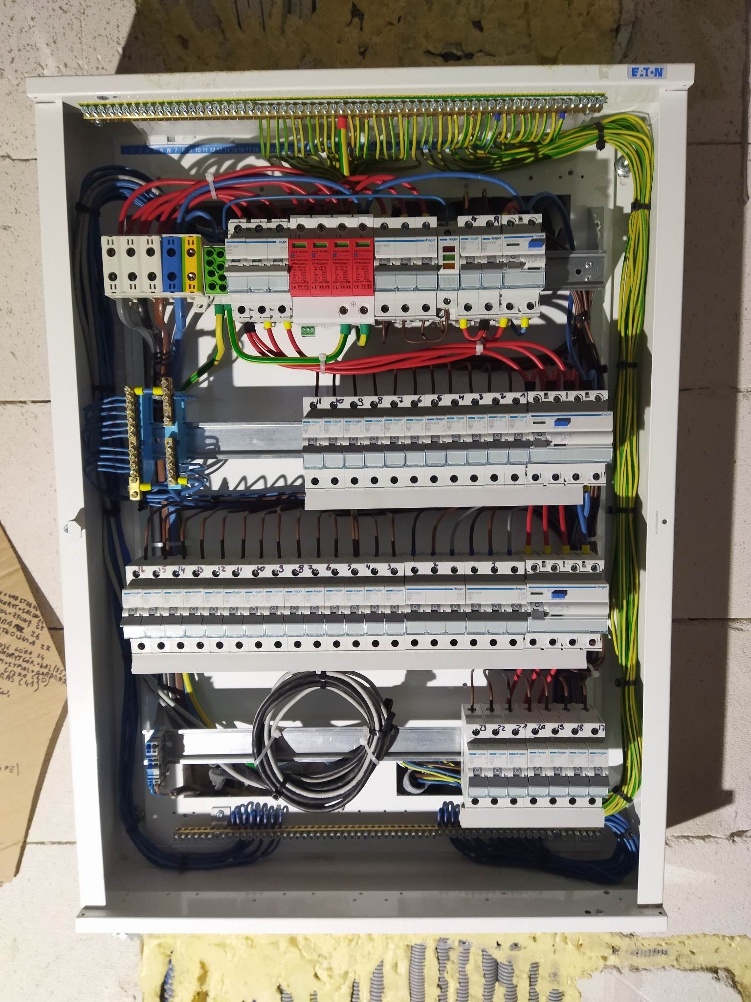 Rozdzielnica elektryczna w białej obudowie, widoczne liczne przewody, bezpieczniki i elementy montażowe. Instalacja w trakcie prac, ściana z bloczków w tle.