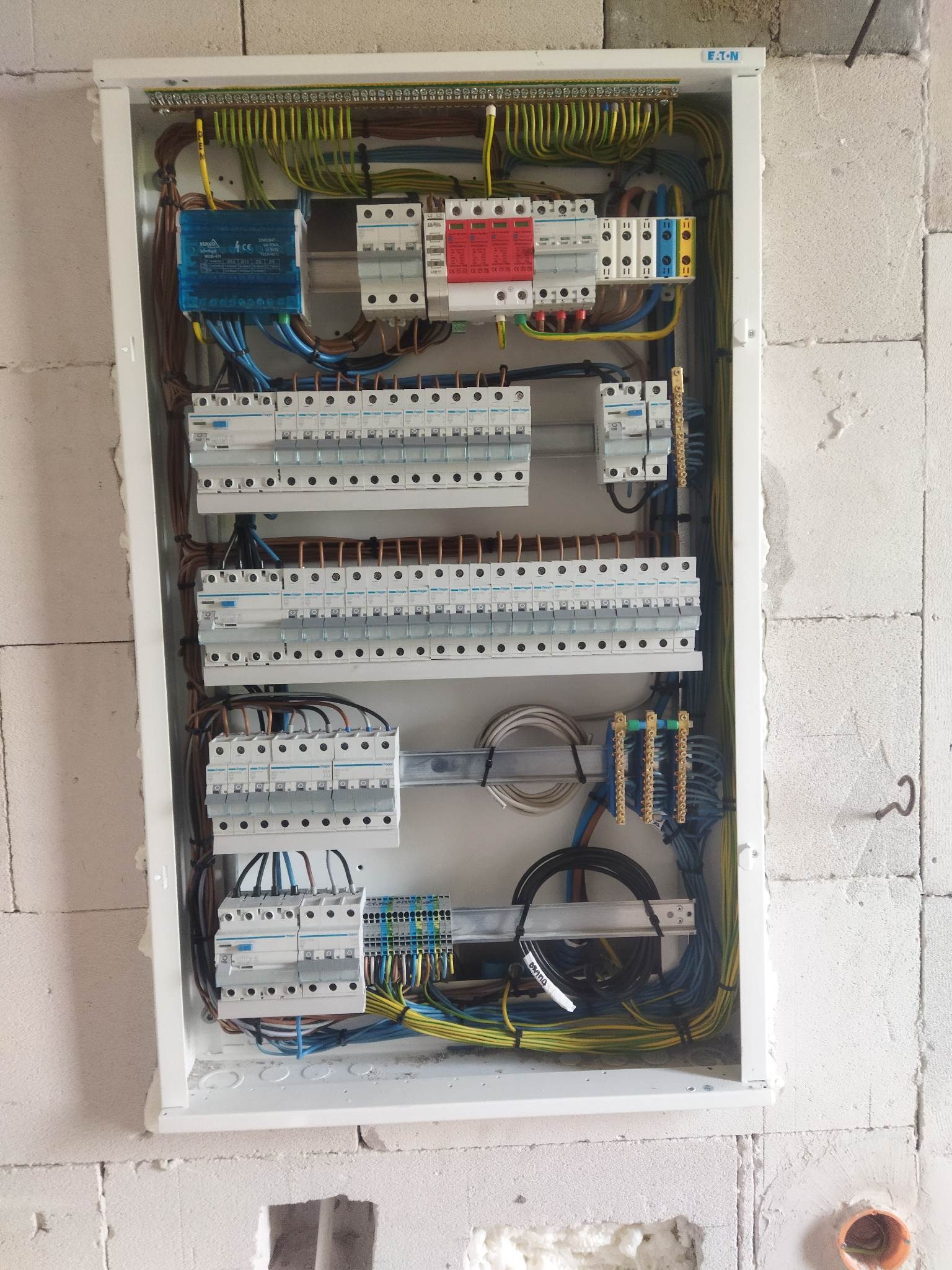 Rozdzielnica elektryczna z licznymi zabezpieczeniami i okablowaniem, zamontowana na ścianie z białych bloczków. Widoczne elementy marki Eaton i Hager.