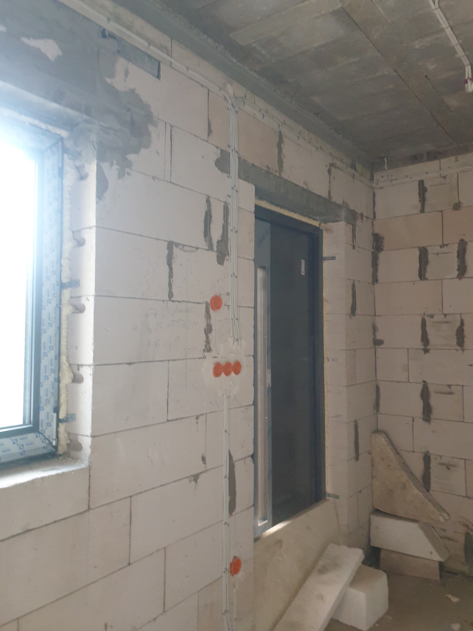 Surowy stan wnętrza z widoczną instalacją elektryczną w peszlach, okno z nieotynkowanym murem, przygotowane podłoże pod tynk. Pomieszczenie w trakcie prac wykończeniowych.