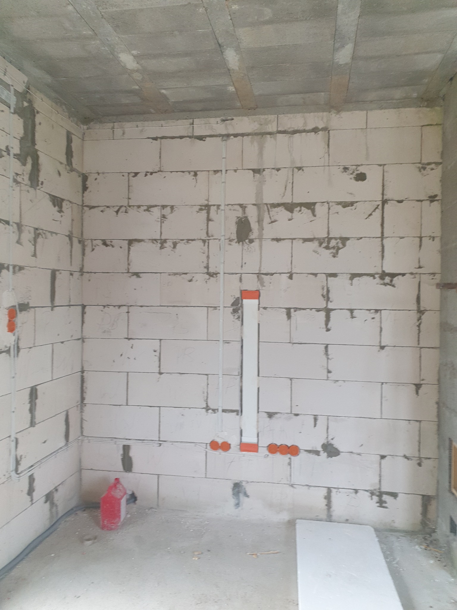 Surowy stan: białe ściany z betonu komórkowego, widoczne przewody elektryczne w peszlach i puszki instalacyjne, sufit betonowy. Wnętrze w trakcie prac wykończeniowych.