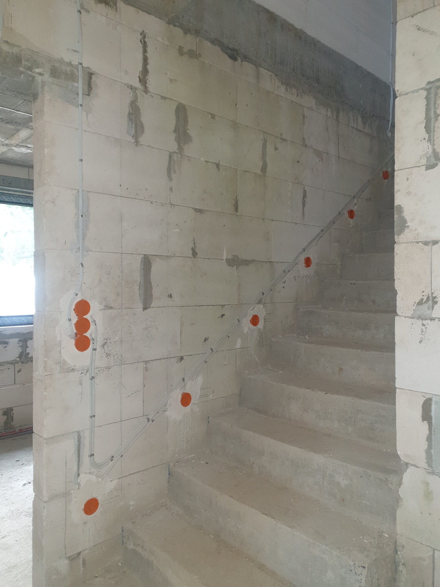 Surowa ściana z betonu komórkowego z poprowadzonymi przewodami elektrycznymi i pomarańczowymi puszkami instalacyjnymi wzdłuż betonowych schodów w surowym stanie.