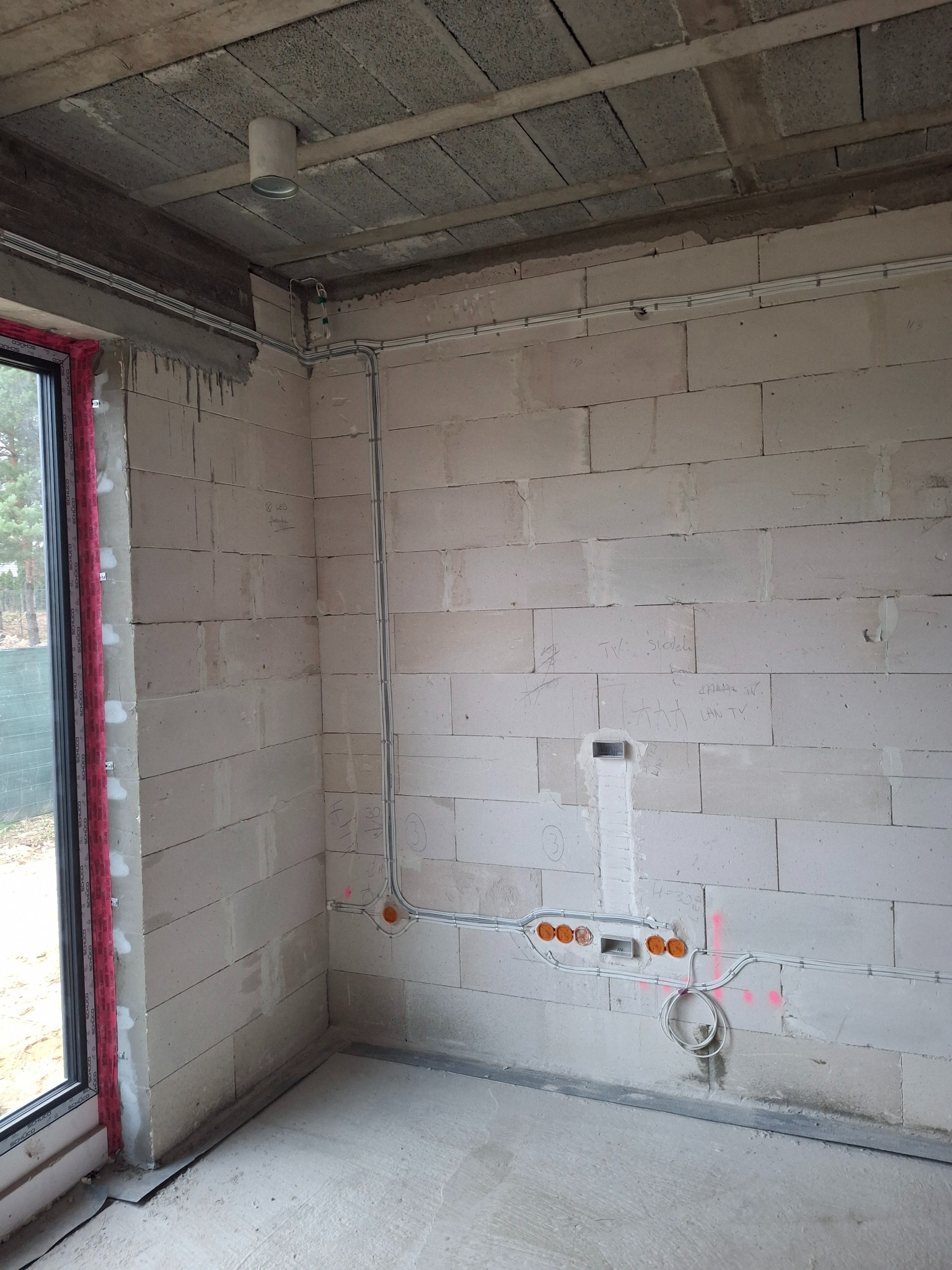 Surowy mur z widoczną instalacją elektryczną: kable w peszlach, puszki podtynkowe. Widoczne oznaczenia na ścianie. Stan surowy budynku, okno z czerwoną folią ochronną.