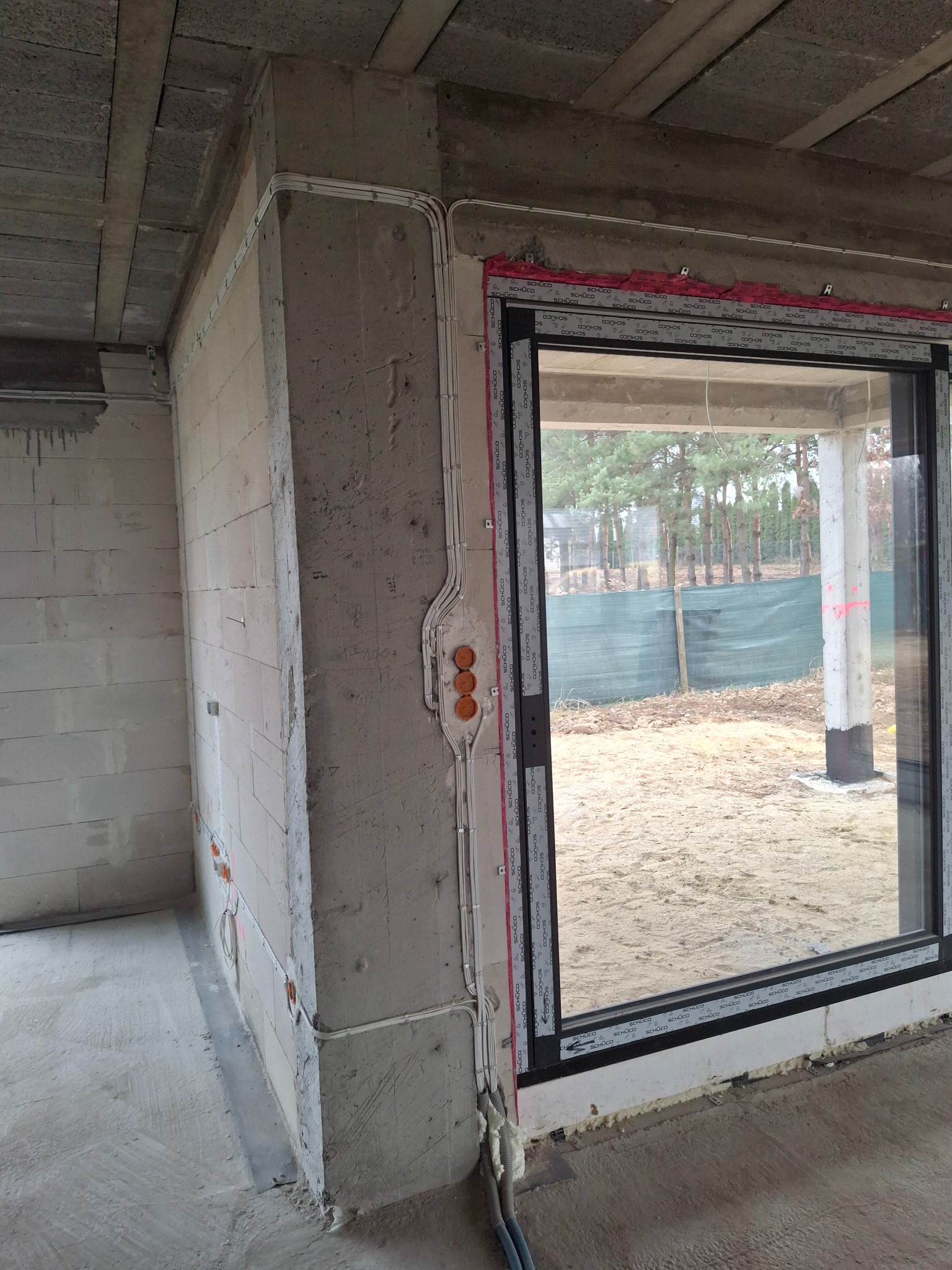 Surowy stan: widok na wnętrze w trakcie prac wykończeniowych, z widocznymi przewodami elektrycznymi na ścianie i oknem z widokiem na zewnątrz.