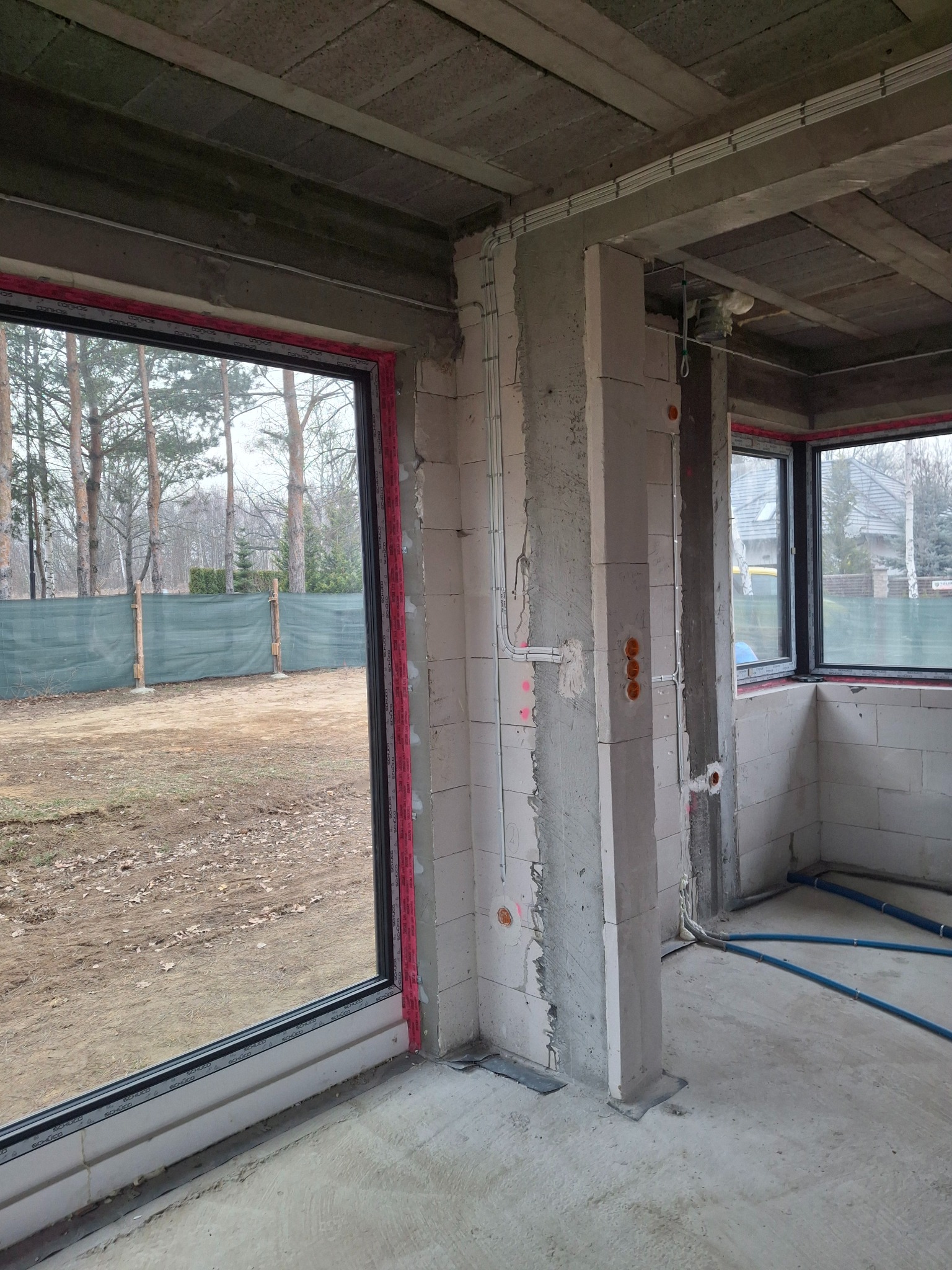 Surowy stan wnętrza domu w budowie z widocznymi przewodami elektrycznymi na ścianach i suficie, okna z czarnymi ramami, betonowy strop.