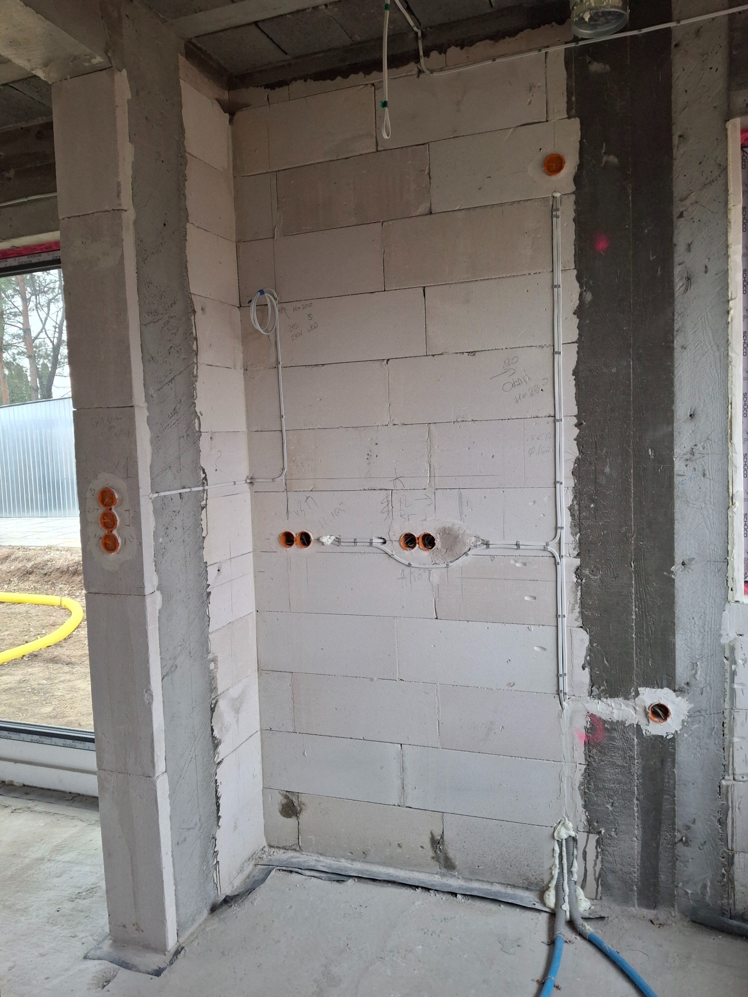 Surowy mur z instalacją elektryczną w trakcie montażu: kable w peszlach, puszki podtynkowe, widoczne oznaczenia na ścianie. Etap prac przed tynkowaniem.