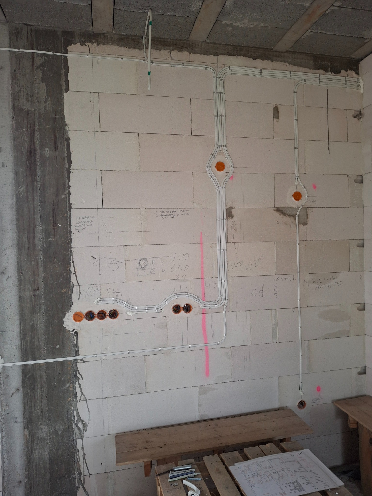 Białe przewody elektryczne ułożone na surowej ścianie z betonu komórkowego, z widocznymi puszkami instalacyjnymi i odręcznymi notatkami technicznymi na ścianie.