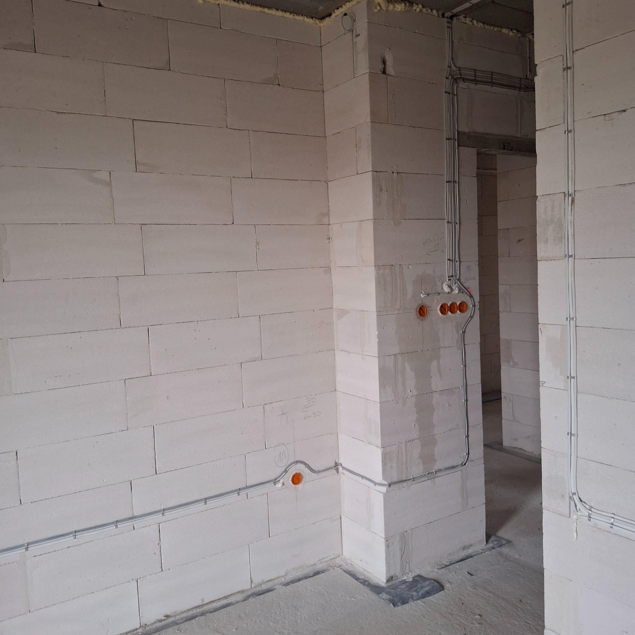 Surowy stan: instalacja elektryczna w peszlach na ścianie z betonu komórkowego, widoczne puszki instalacyjne. Przewody prowadzone wzdłuż ścian, w tle widoczne przejście.