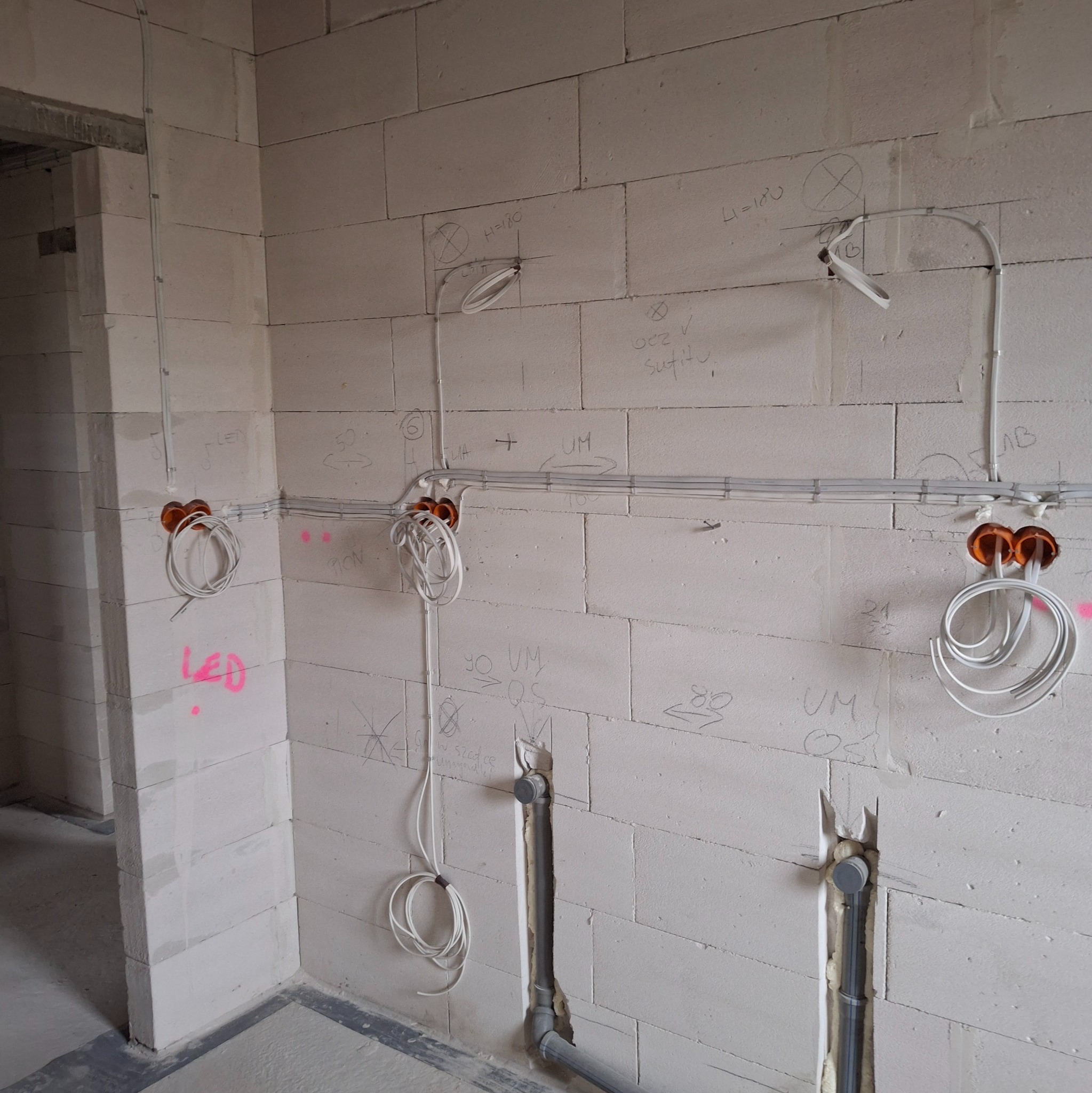 Surowy stan: instalacja elektryczna w ścianach z betonu komórkowego, widoczne kable, puszki i rury. Ściany z odręcznymi notatkami i oznaczeniami.
