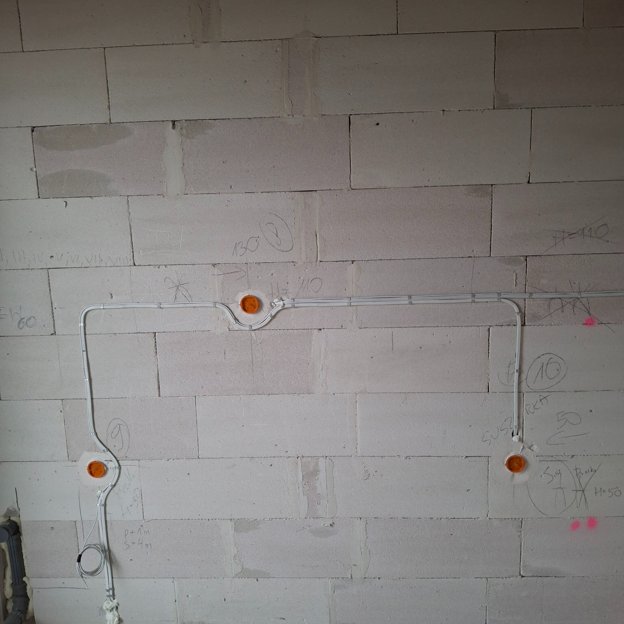 Białe przewody elektryczne ułożone na szarej ścianie z betonu komórkowego, z pomarańczowymi puszkami instalacyjnymi i odręcznymi notatkami na ścianie.