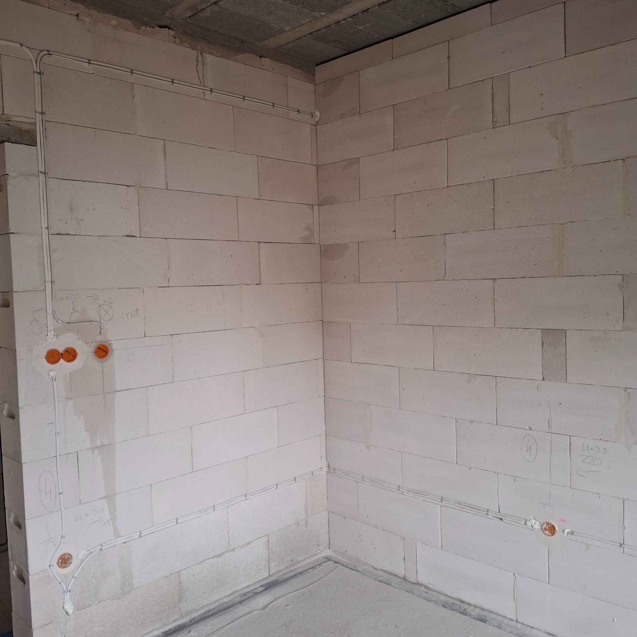 Surowa ściana z betonu komórkowego z poprowadzonymi przewodami elektrycznymi i puszkami instalacyjnymi. Widoczne odręczne notatki na ścianie.