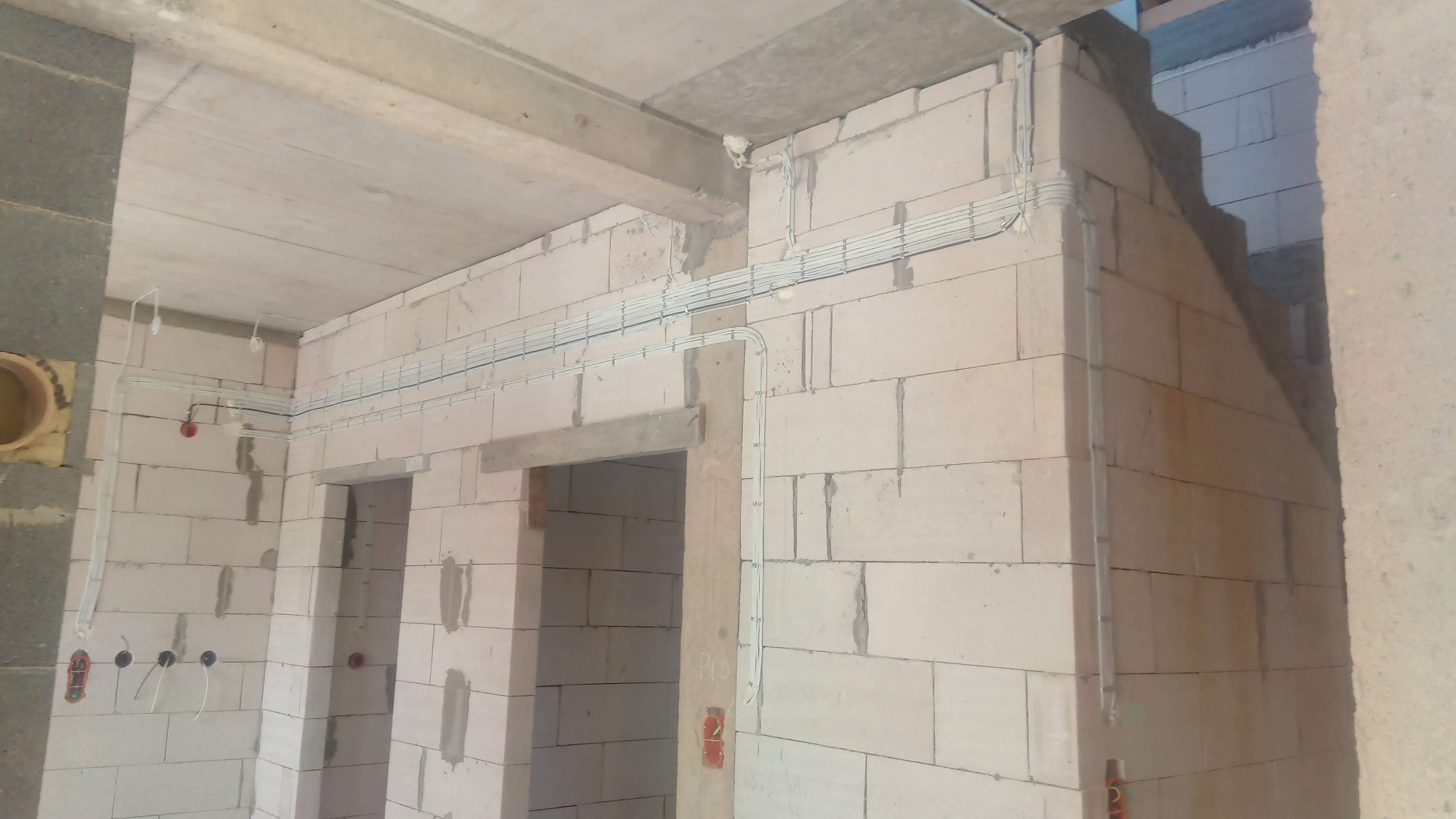 Surowe wnętrze budynku w trakcie prac instalacyjnych: widoczne przewody elektryczne prowadzone po ścianach z bloczków betonowych oraz otwory pod gniazdka i włączniki.