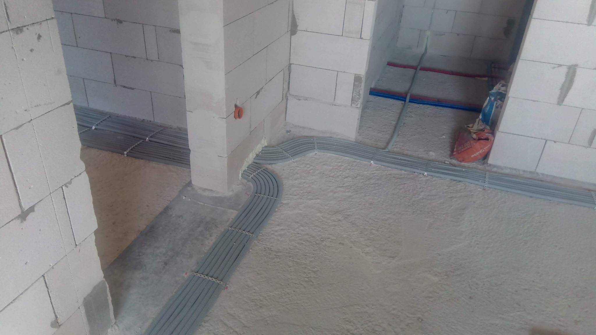 Rury instalacyjne elektryczne ułożone wzdłuż ściany z bloczków betonowych w surowym wnętrzu, zabezpieczone metalowymi uchwytami.