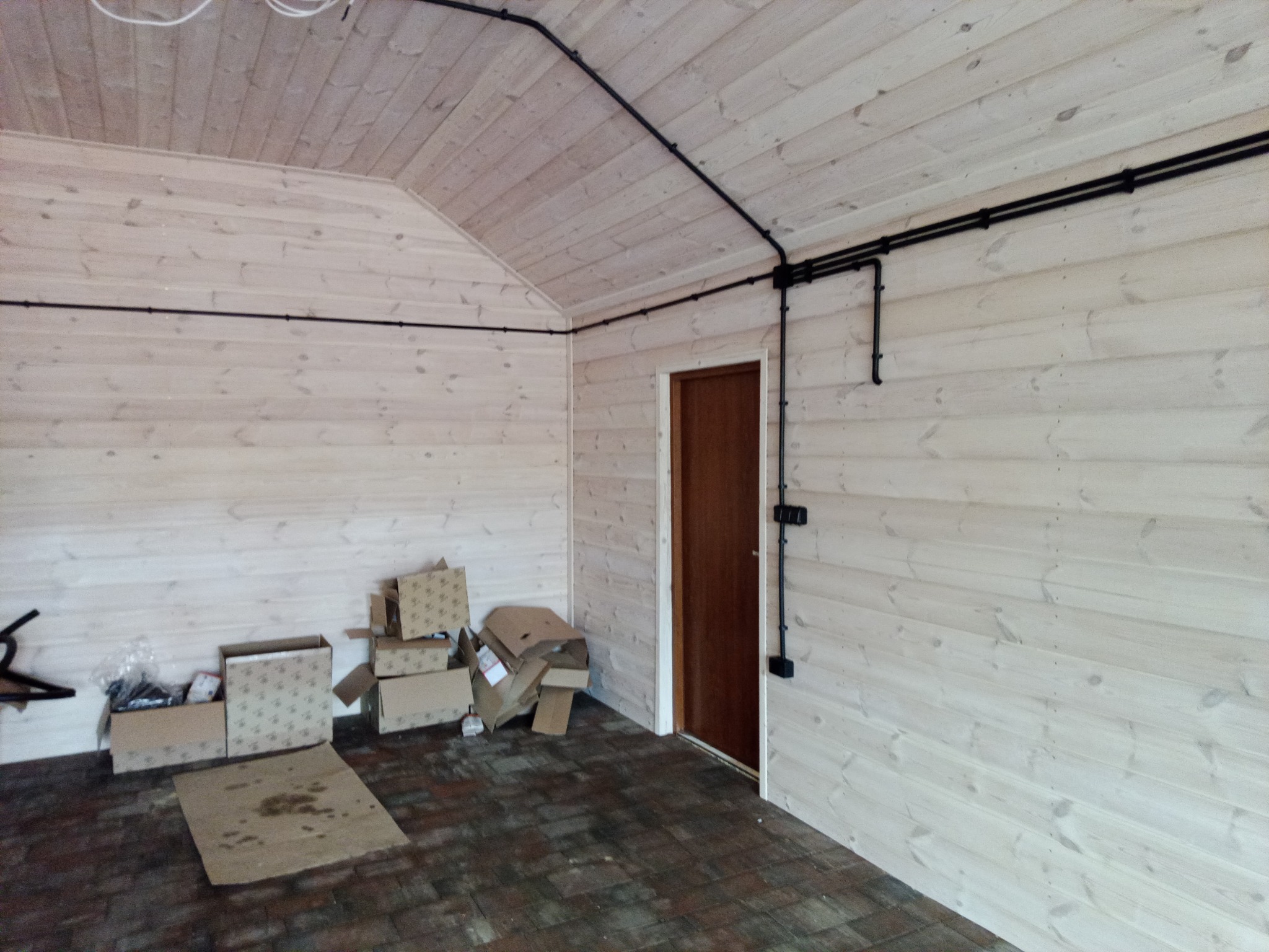 Wnętrze wykończonego pomieszczenia z drewnianą boazerią, gdzie po ścianach i suficie poprowadzone są czarne rury instalacji elektrycznej; widoczne puszki instalacyjne, a na podłodze leżą kartony.