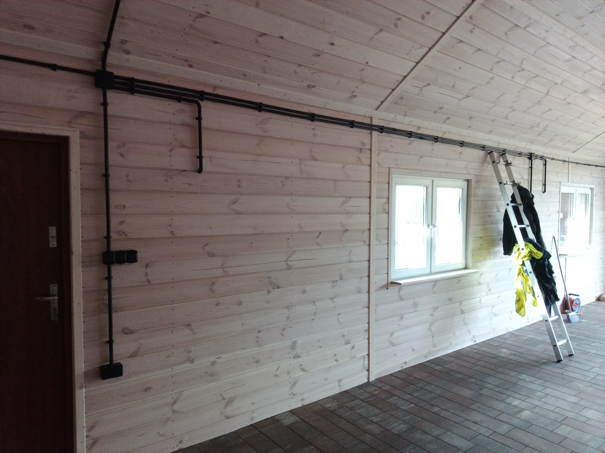 Czarne rury instalacji elektrycznej poprowadzone po ścianie i suficie wykończonym drewnem w jasnym pomieszczeniu. Przy ścianie stoi drabina z roboczymi ubraniami.