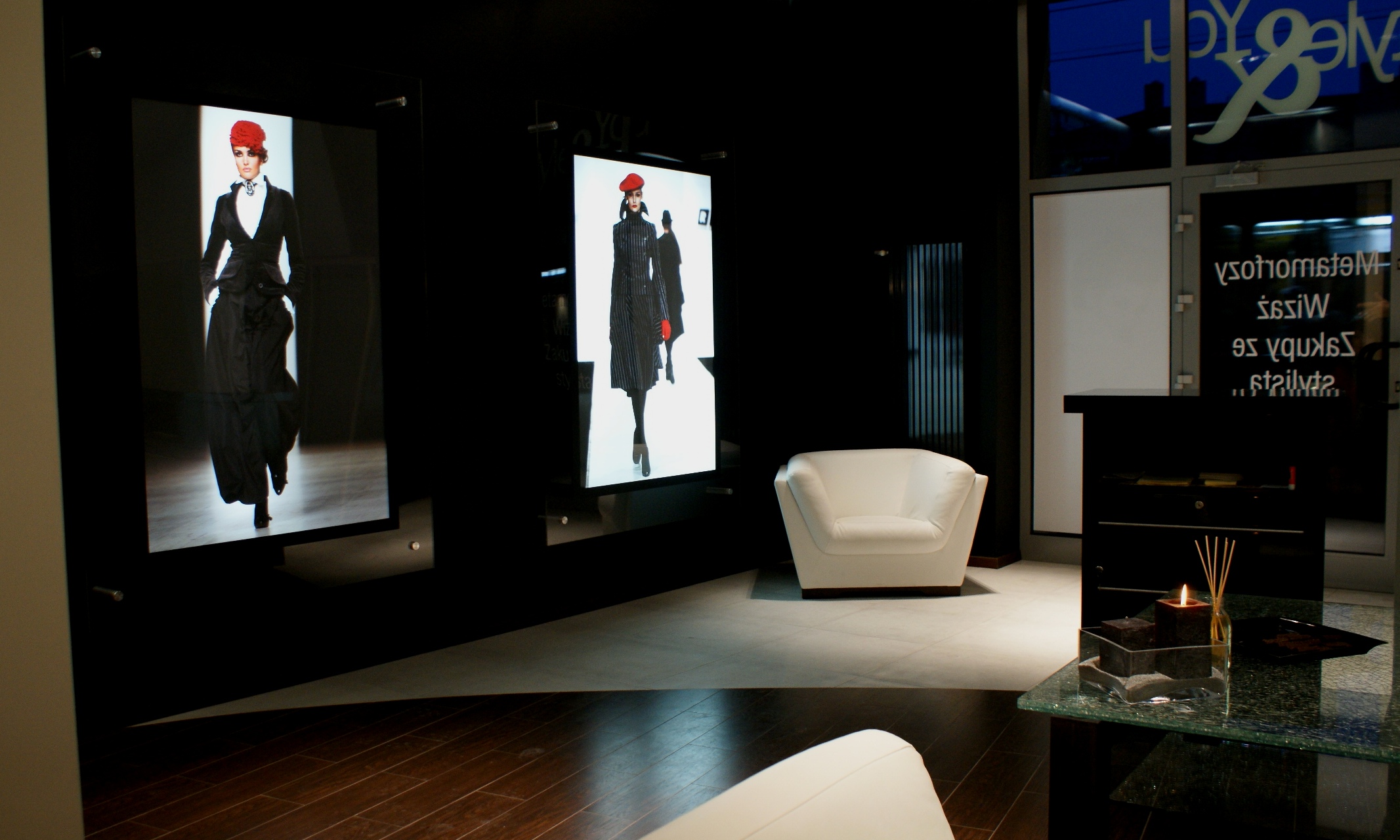 Nowoczesne wnętrze butiku z dwoma podświetlanymi ekranami przedstawiającymi modelki w czarnych strojach i czerwonych beretach, biały fotel i stolik kawowy ze świecą.
