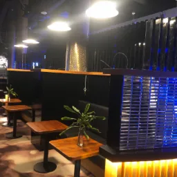 Nowoczesne wnętrze restauracji z ciemnymi lożami, drewnianymi stolikami, roślinami w szklanych wazonach i podświetlanymi elementami dekoracyjnymi w odcieniach niebieskiego i żółtego.