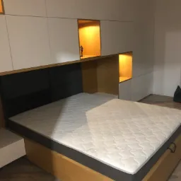 Nowoczesna sypialnia z łóżkiem na drewnianej ramie, wezgłowiem z szarych paneli i zabudową meblową z białymi szafkami oraz podświetlanymi wnękami z drewnianym wykończeniem, w jednej z wnęk stoi...