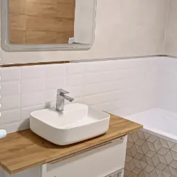 Nowoczesna, kwadratowa umywalka nablatowa z chromowaną baterią na drewnianym blacie szafki w łazience we Wrocławiu, z widocznym fragmentem wanny obłożonej geometryczną mozaiką i podświetlanym lustrem.