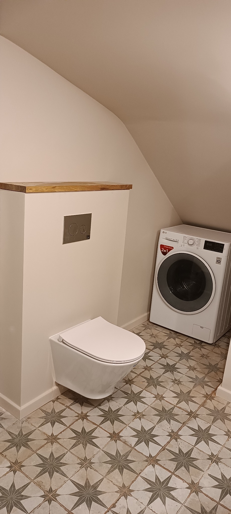Nowoczesna łazienka na poddaszu ze ścianą zabudowaną drewnianym blatem nad toaletą podwieszaną i pralką LG na podłodze z wzorzystych płytek.