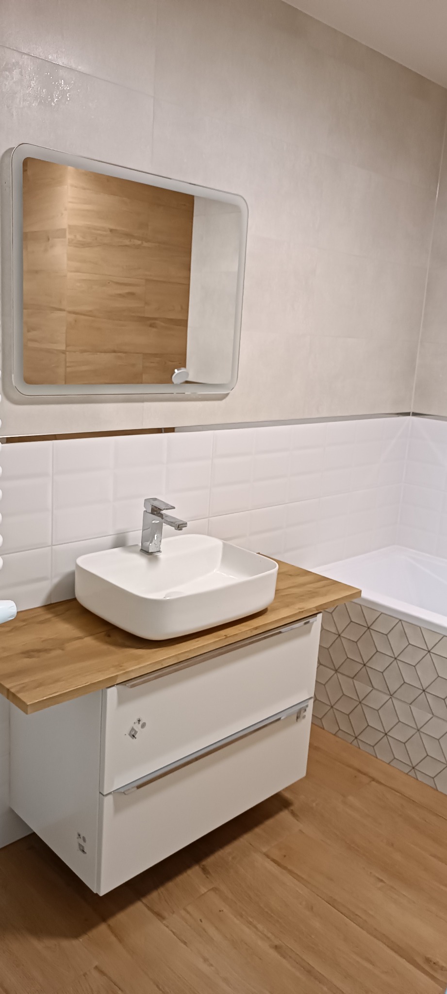 Nowoczesna, kwadratowa umywalka nablatowa z chromowaną baterią na drewnianym blacie szafki w łazience we Wrocławiu, z widocznym fragmentem wanny obłożonej geometryczną mozaiką i podświetlanym lustrem.