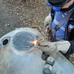 Spawanie palnikiem gazowym metalowej konstrukcji na zewnątrz, widoczne iskry, osłona spawalnicza na twarzy spawacza, obok leżą trzy metalowe tuleje z gwintem.