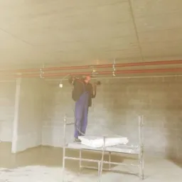 Instalator w kombinezonie roboczym na rusztowaniu spawa rury instalacji pod sufitem w surowym pomieszczeniu z betonowymi ścianami.