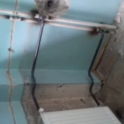 Fragment instalacji grzewczej z rurami przy ścianie pomalowanej na błękitno i fragmentem grzejnika, widoczne ślady po pracach remontowych na podłodze.