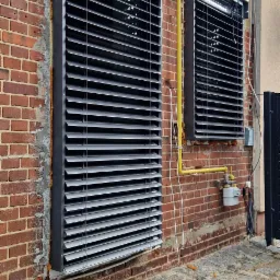 Dwa okna z szarymi żaluzjami fasadowymi zamontowane na ceglanej ścianie budynku z widoczną instalacją gazową