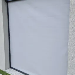 Zamknięta, szara roleta zewnętrzna z czarną ramą, zamontowana na elewacji domu, z widocznym fragmentem sztucznej trawy i lampy elewacyjnej.